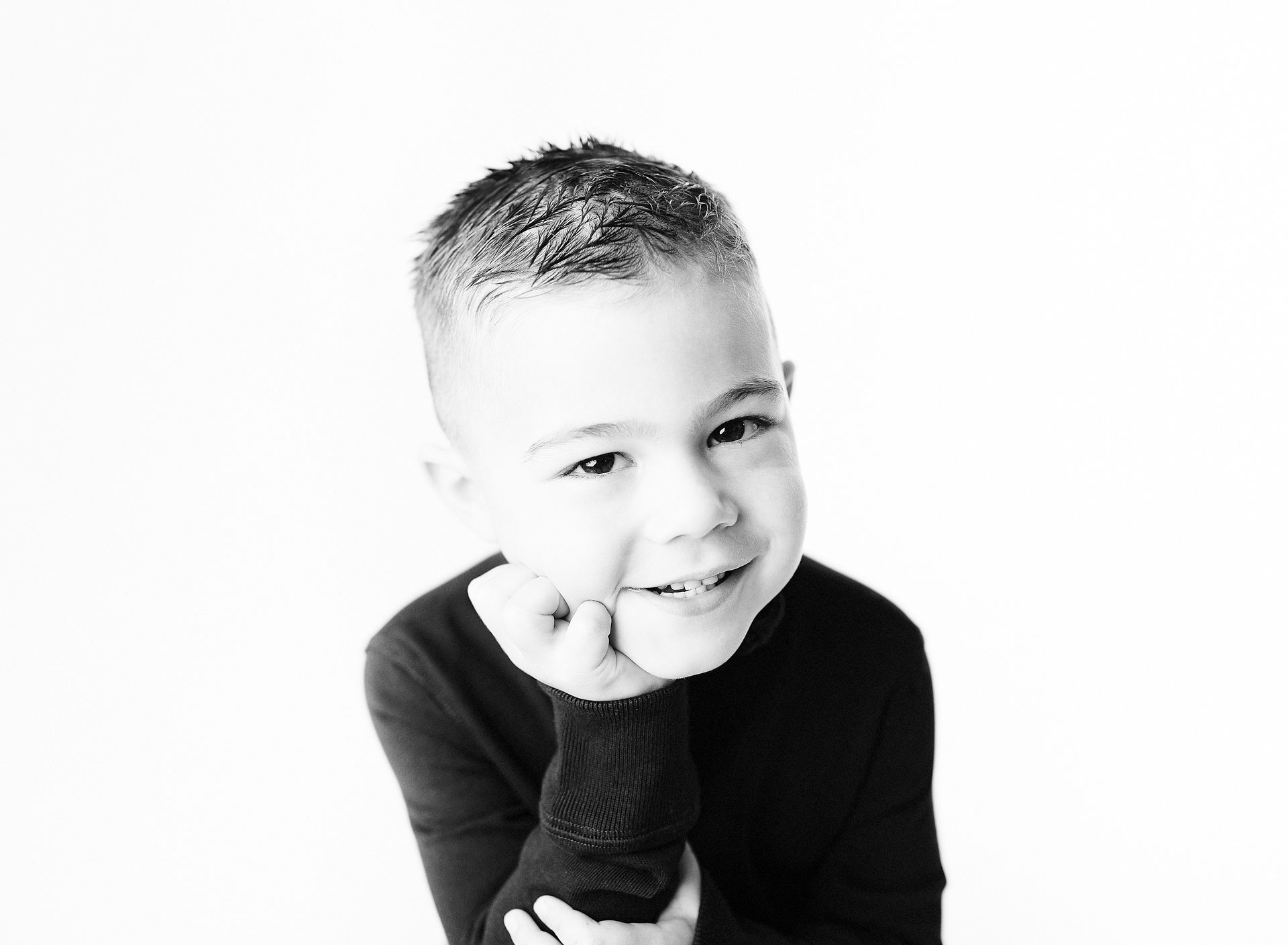 Boy smiling, resting chin on hand against a white backdrop, wearing a dark sweater.