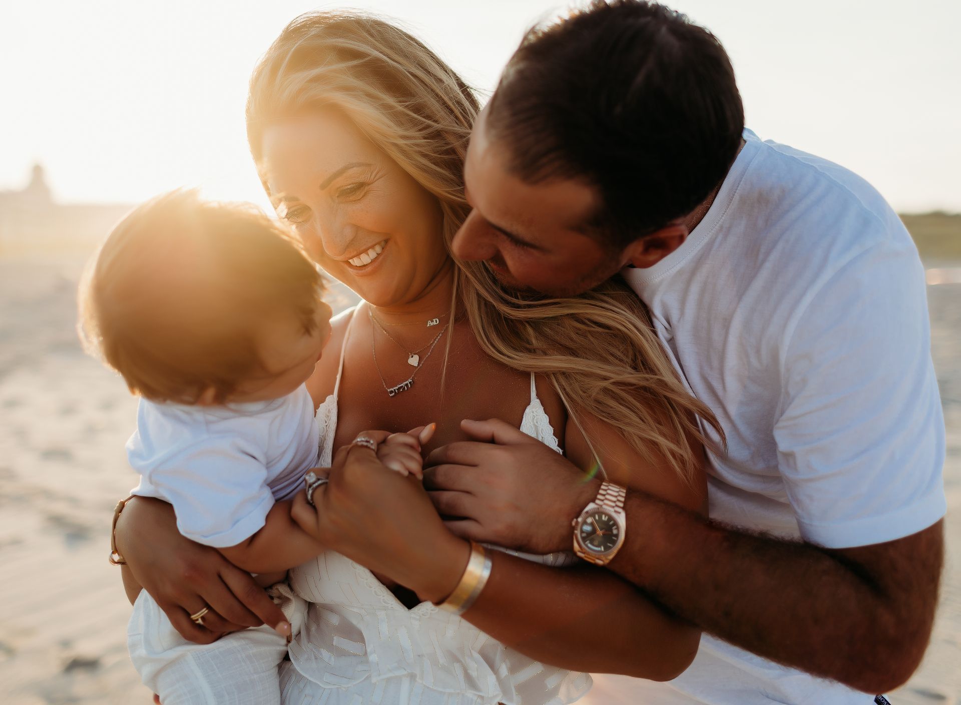 Family hugging on a beach at sunset; a person kisses another's cheek; happy smiles and warm lighting.
