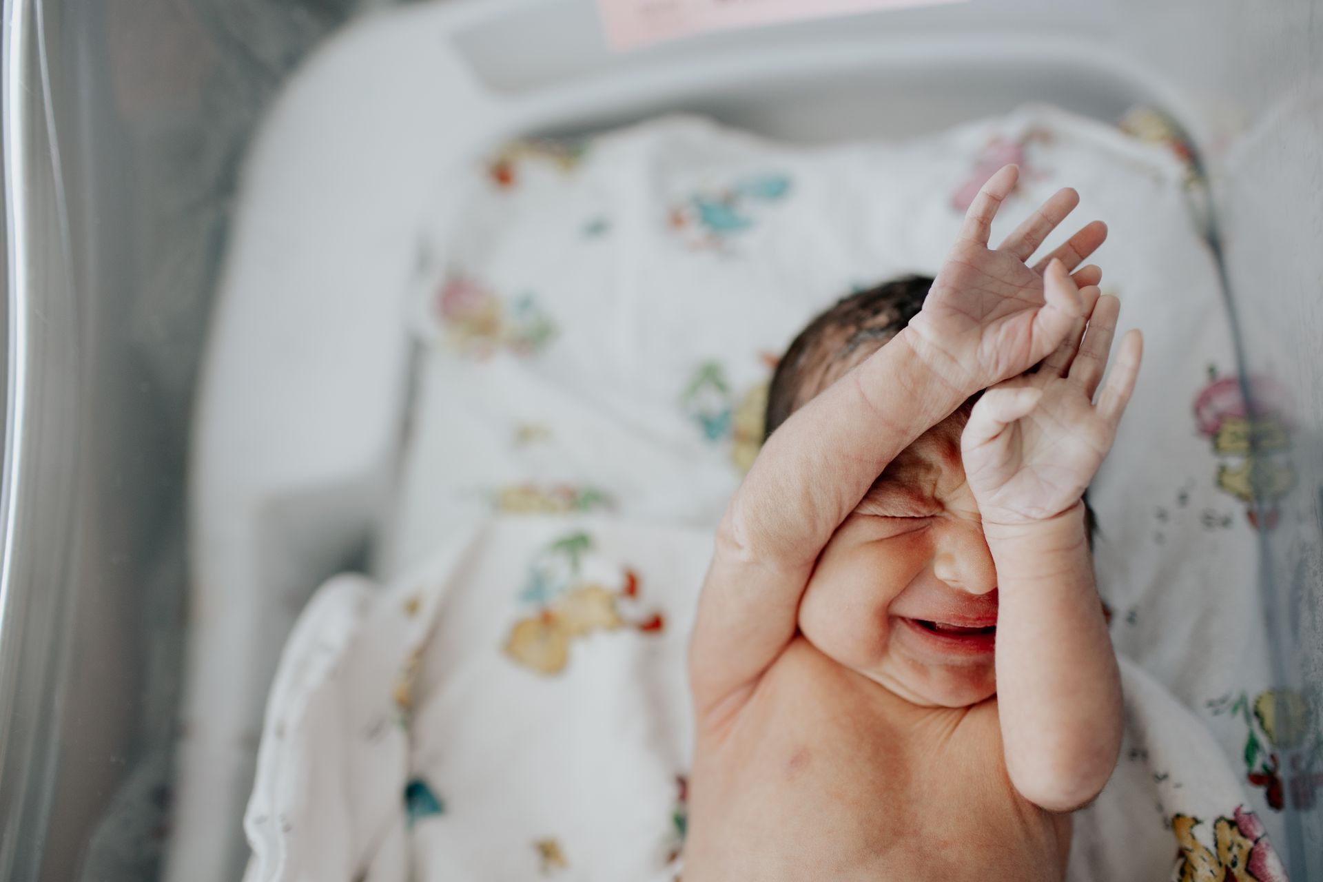 Newborn baby in a hospital bassinet, raising arms, with a crying expression.