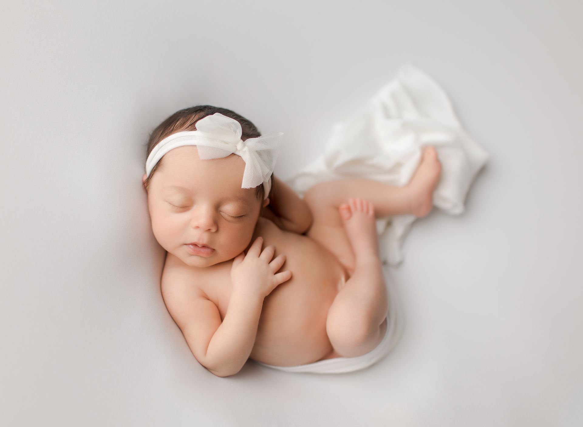 Newborn baby sleeping on white backdrop, wearing white headband.