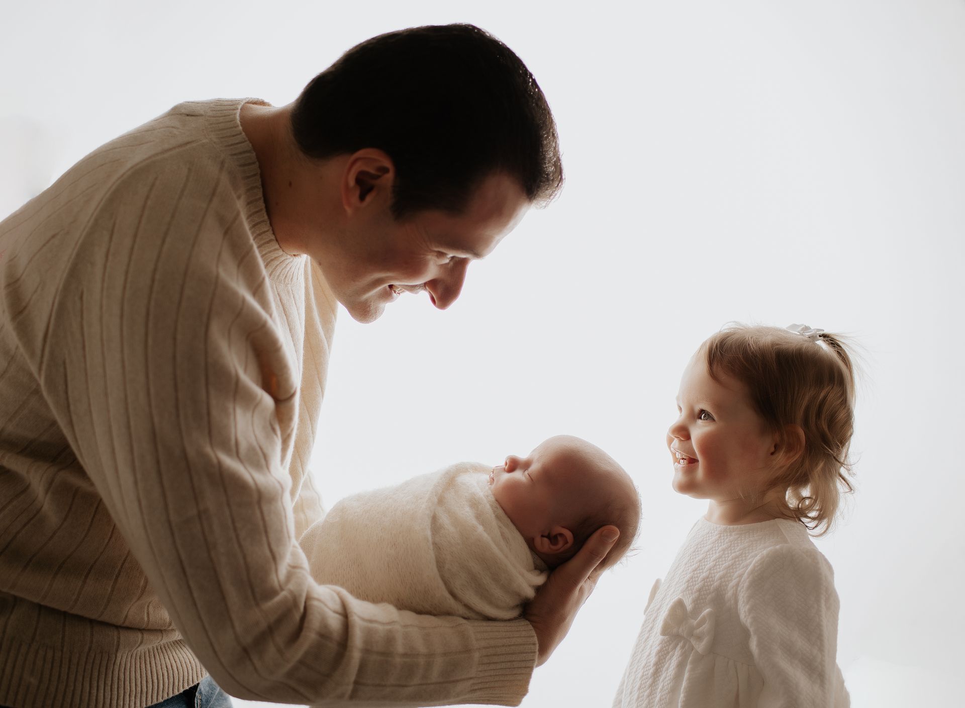 Father holding newborn baby, smiling at toddler daughter; bright white background.