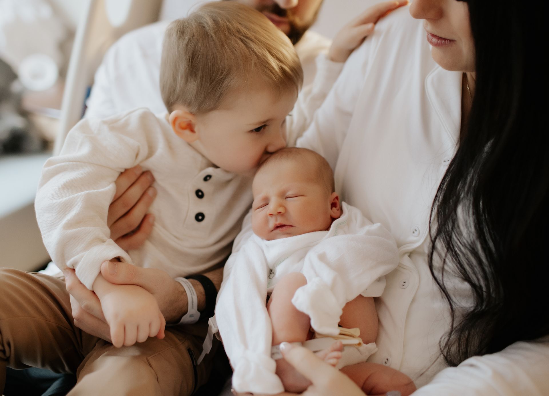 Family with newborn; older sibling kisses baby's head. All are dressed in white indoors.