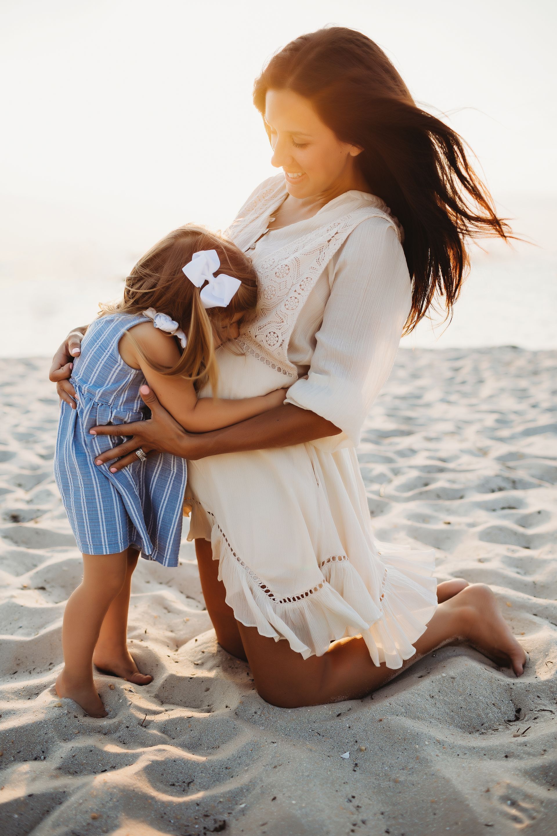 Pregnant woman kneeling on beach, hugging small child's head against her belly. Warm sunlight.