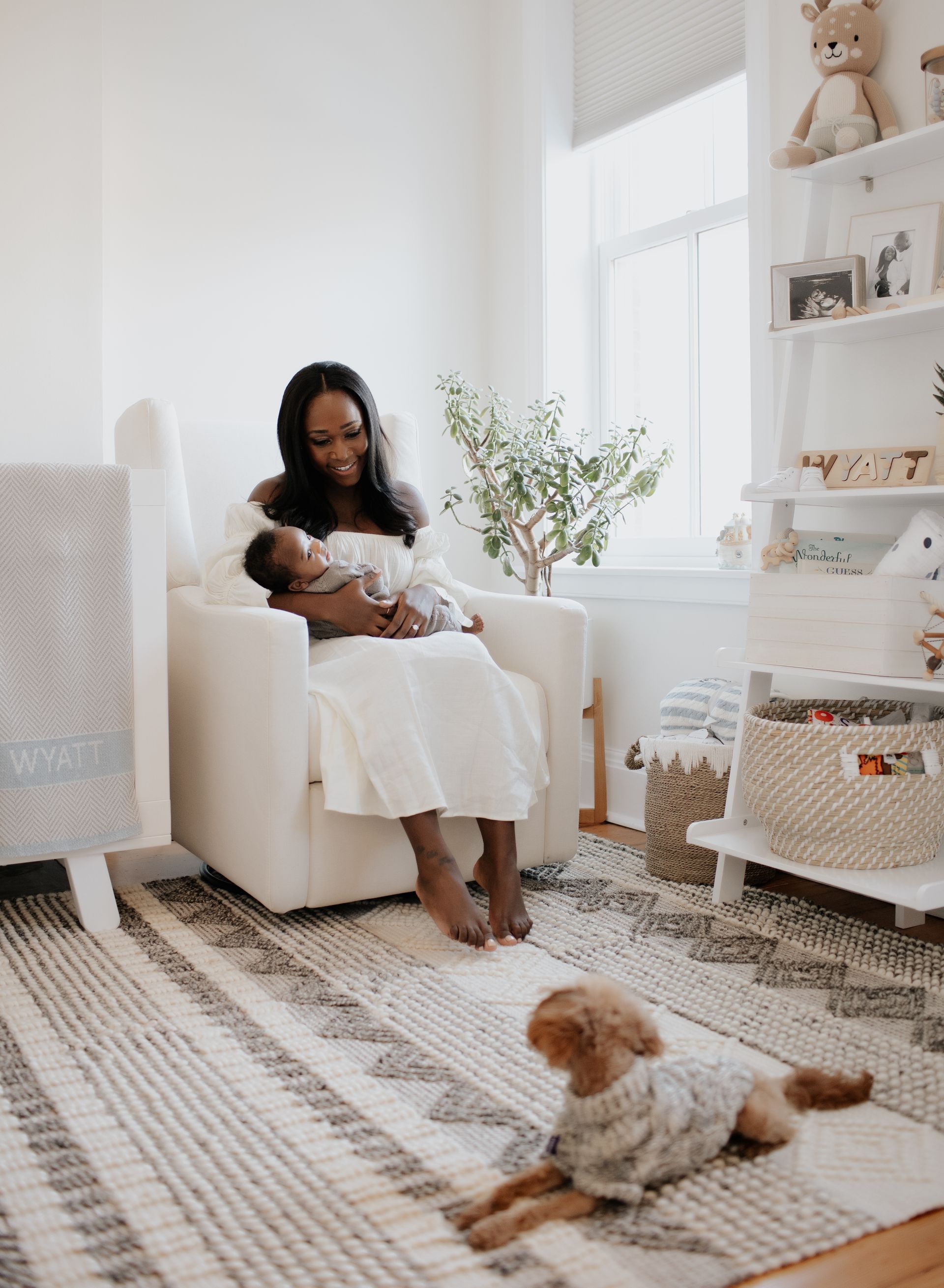 A woman is sitting in a chair holding a baby and a dog is sitting next to her.