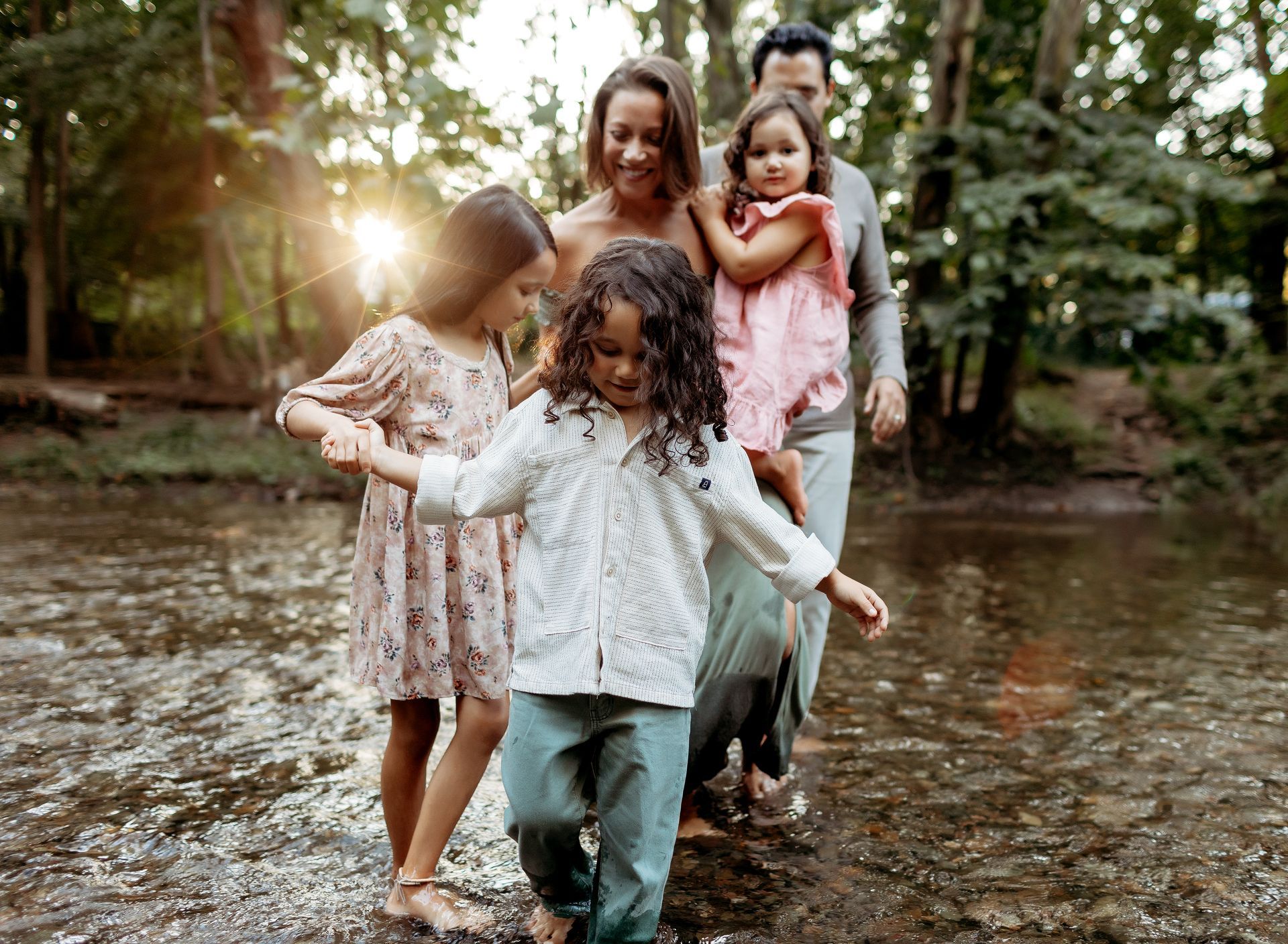 Family wading in a creek, holding hands, smiling. Sunlight streams through trees in background.
