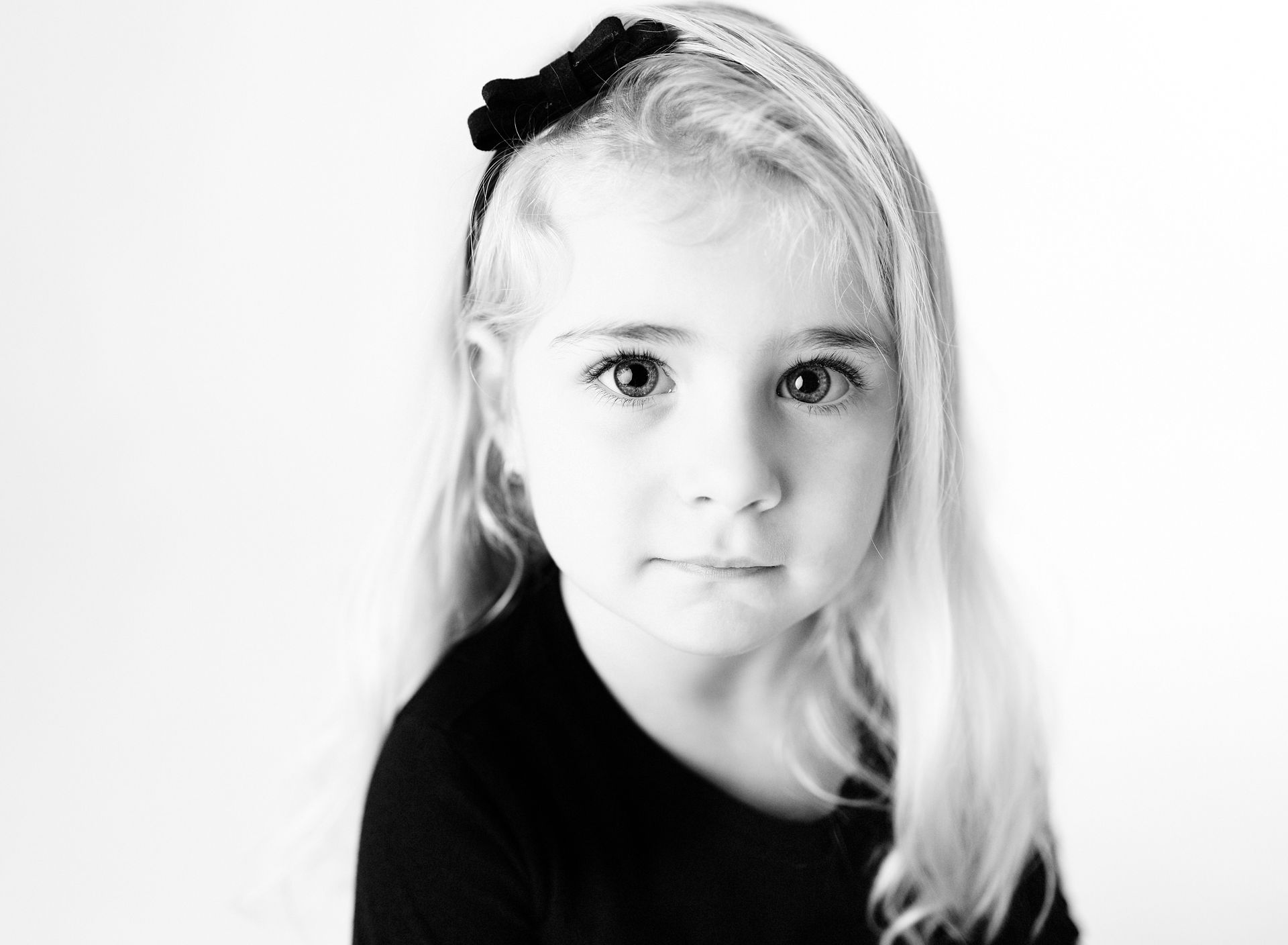 Blond child with a black bow in her hair, looking at the viewer. Black shirt, against a white background.