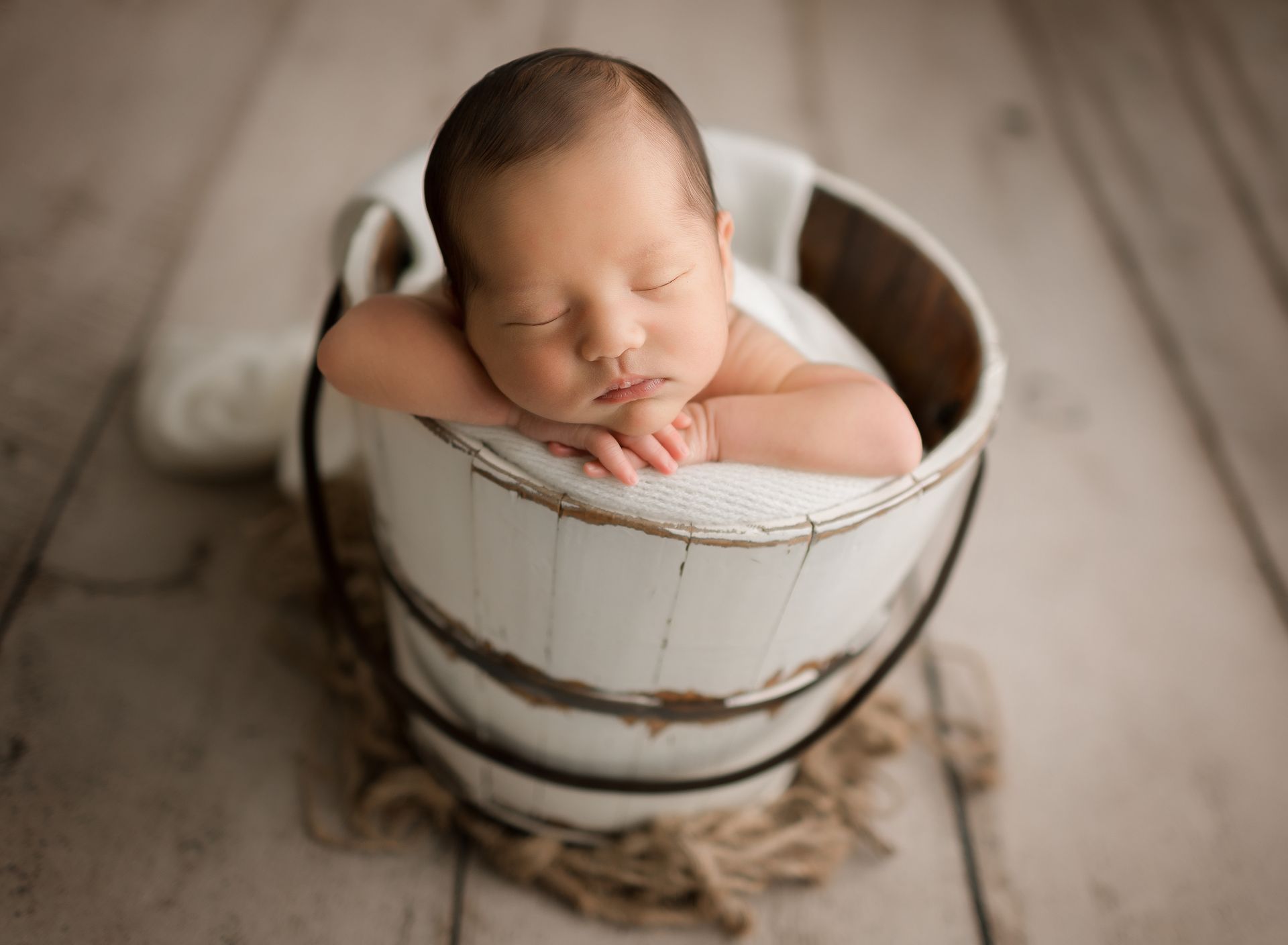 Newborn sleeps peacefully in a white wooden bucket, arms crossed, on a wood floor.