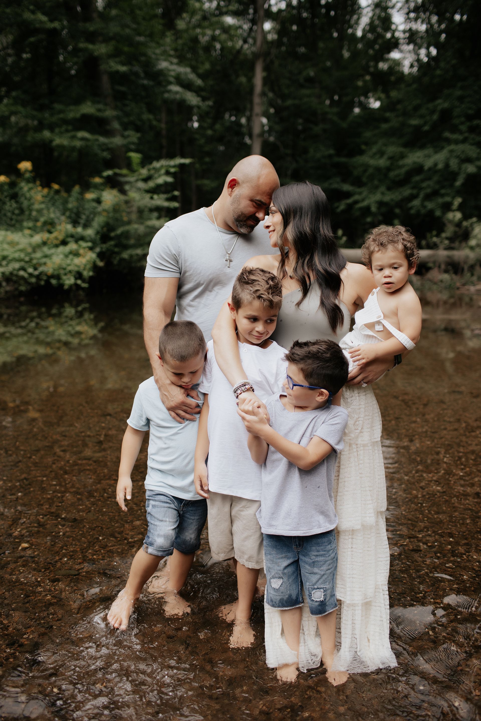 Family of seven standing in shallow water, embracing. Two parents, five children in casual wear, forest setting.