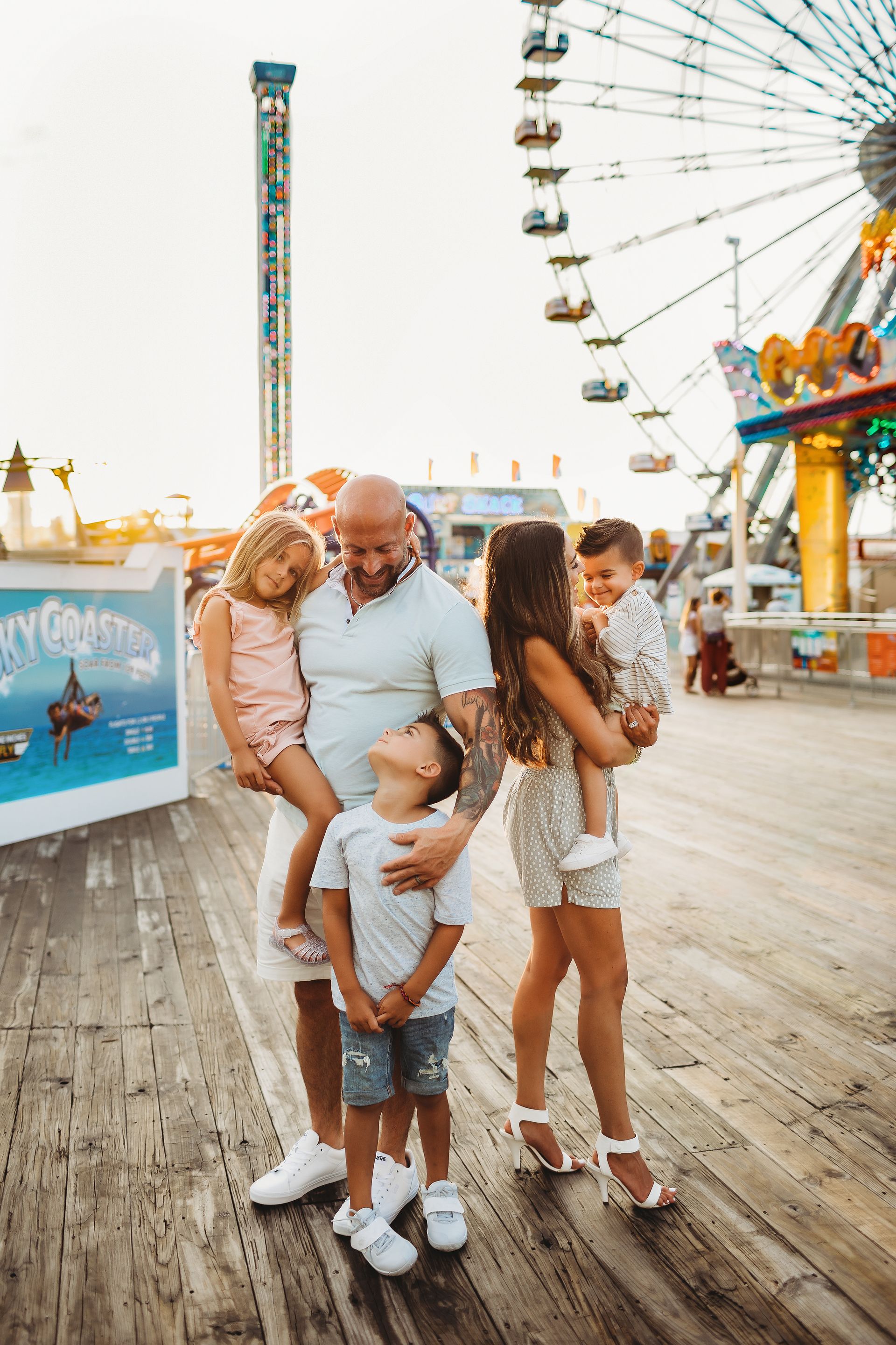 Family posing on boardwalk in front of Ferris wheel. Father holds two children, mother holds one.