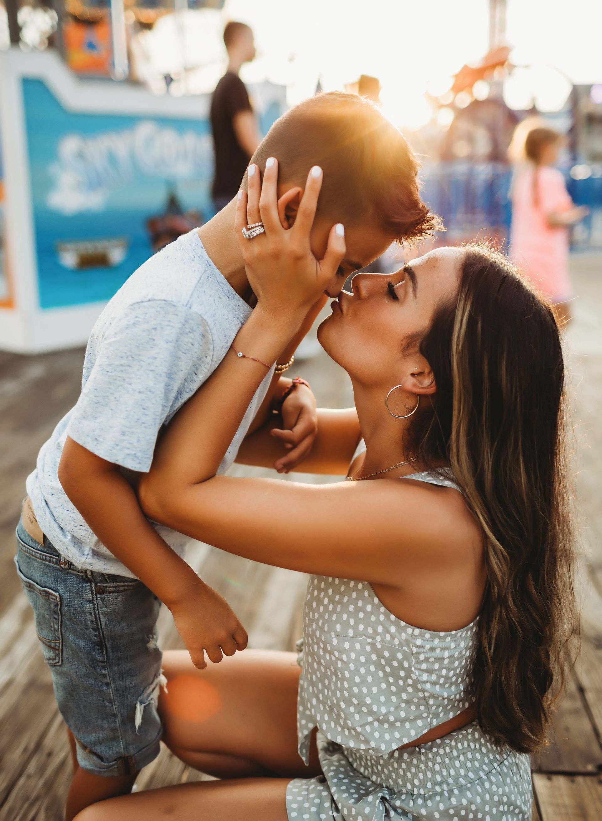 Woman kissing a child on the pier at sunset; child is wearing a blue shirt and denim shorts.