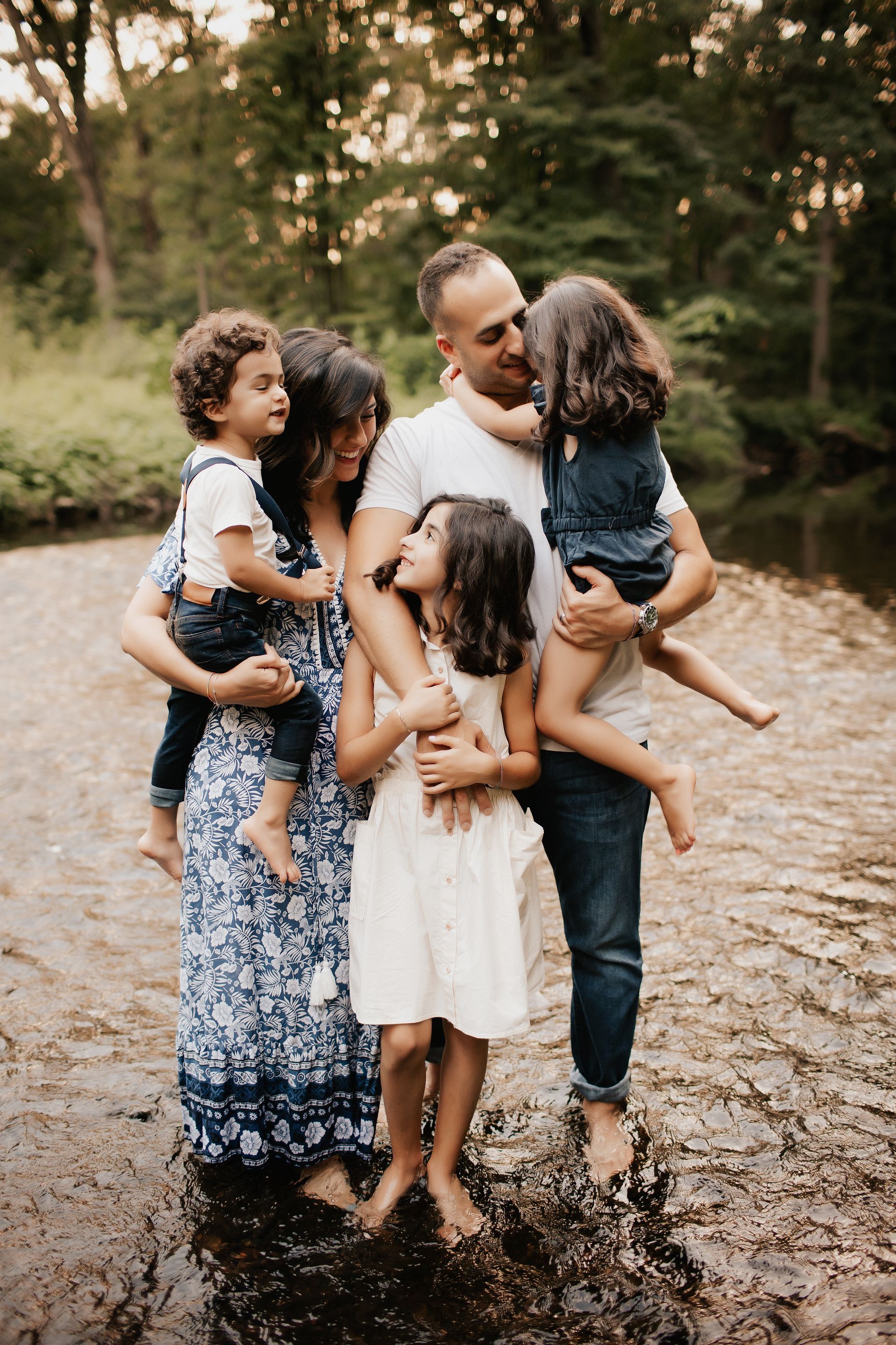 Family wading in a stream. Parents hold children, smiling. Water reflects trees.