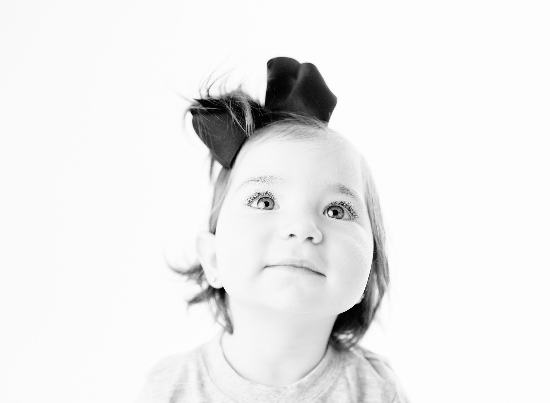 Child with a large bow smiles, looking up. Black and white photo.