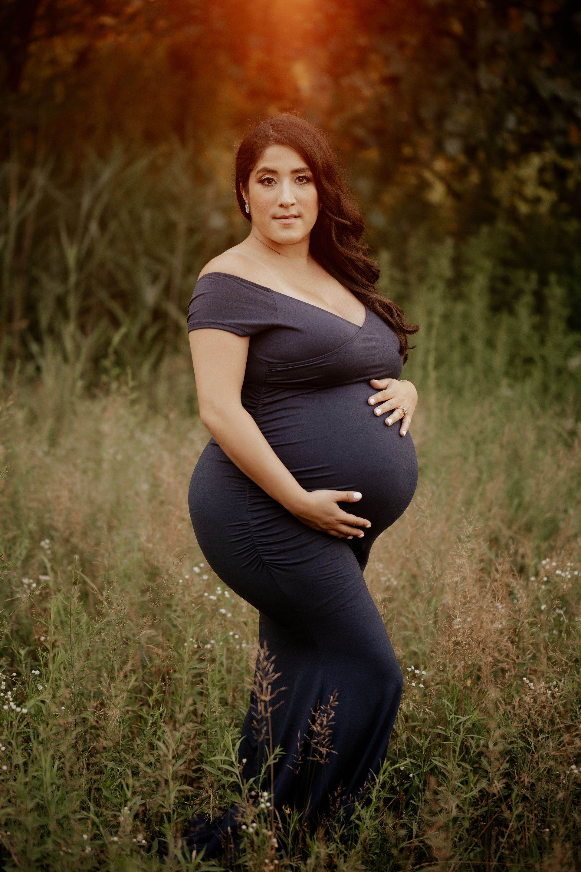 Pregnant person in navy dress, holding belly, outdoors in a field of tall grass, warm sunlight.