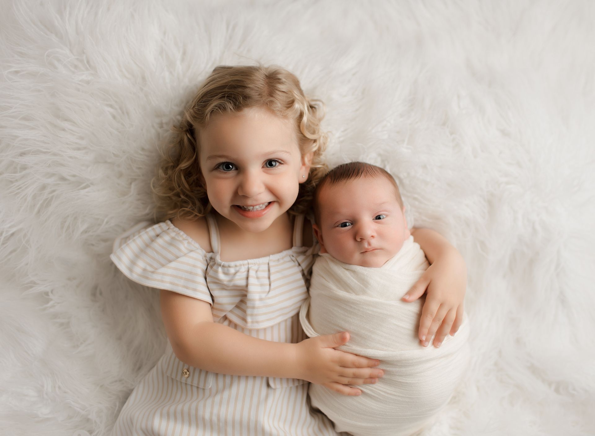 Girl smiling, holding a swaddled infant on a white furry blanket.