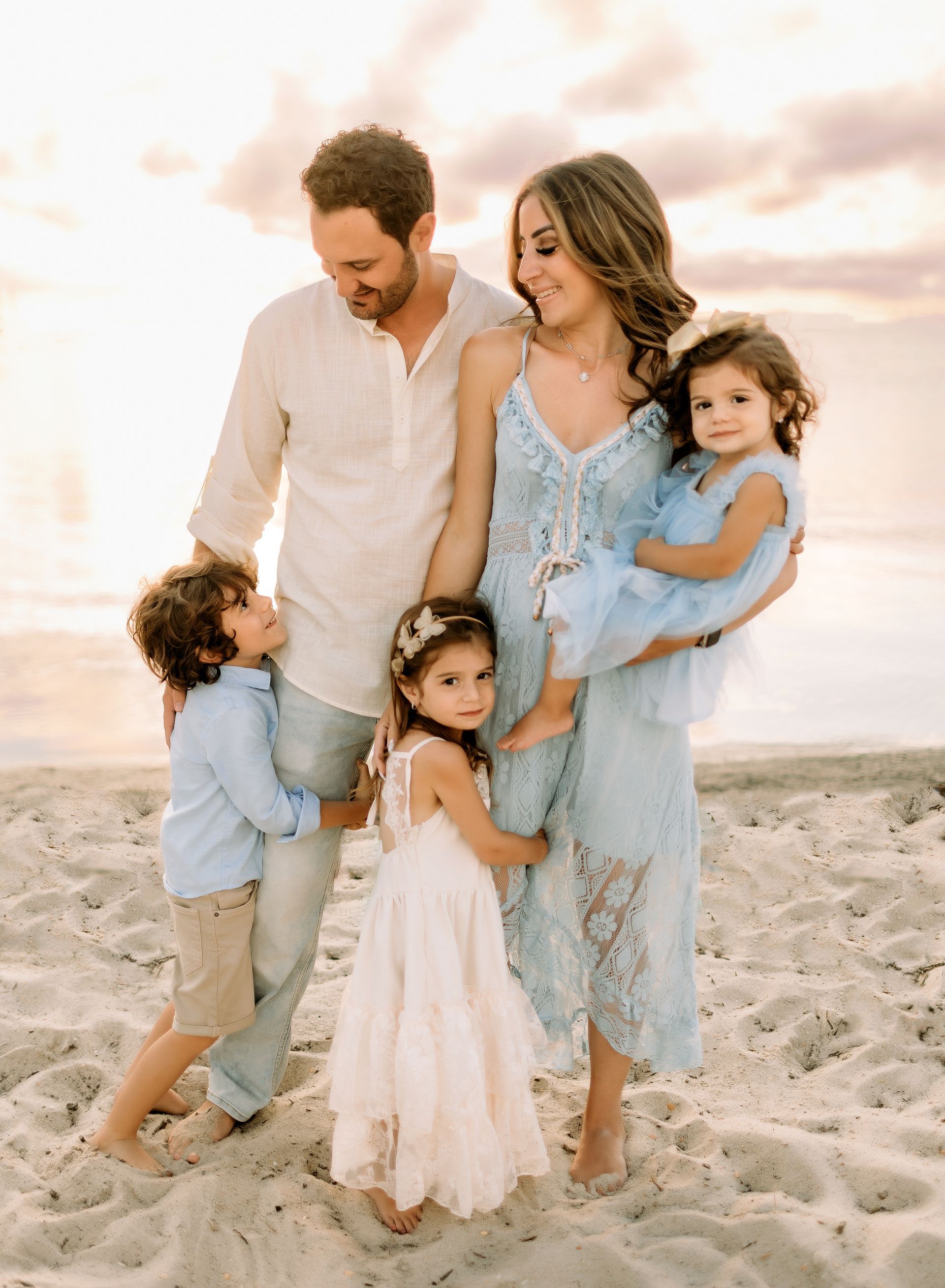 Family poses on beach at sunset: parents with three children in light-colored clothes.