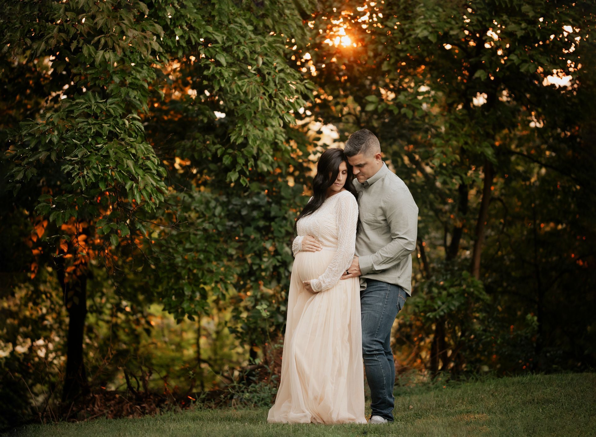 Pregnant couple in embrace, outdoors in front of a tree. Warm sunset lighting.