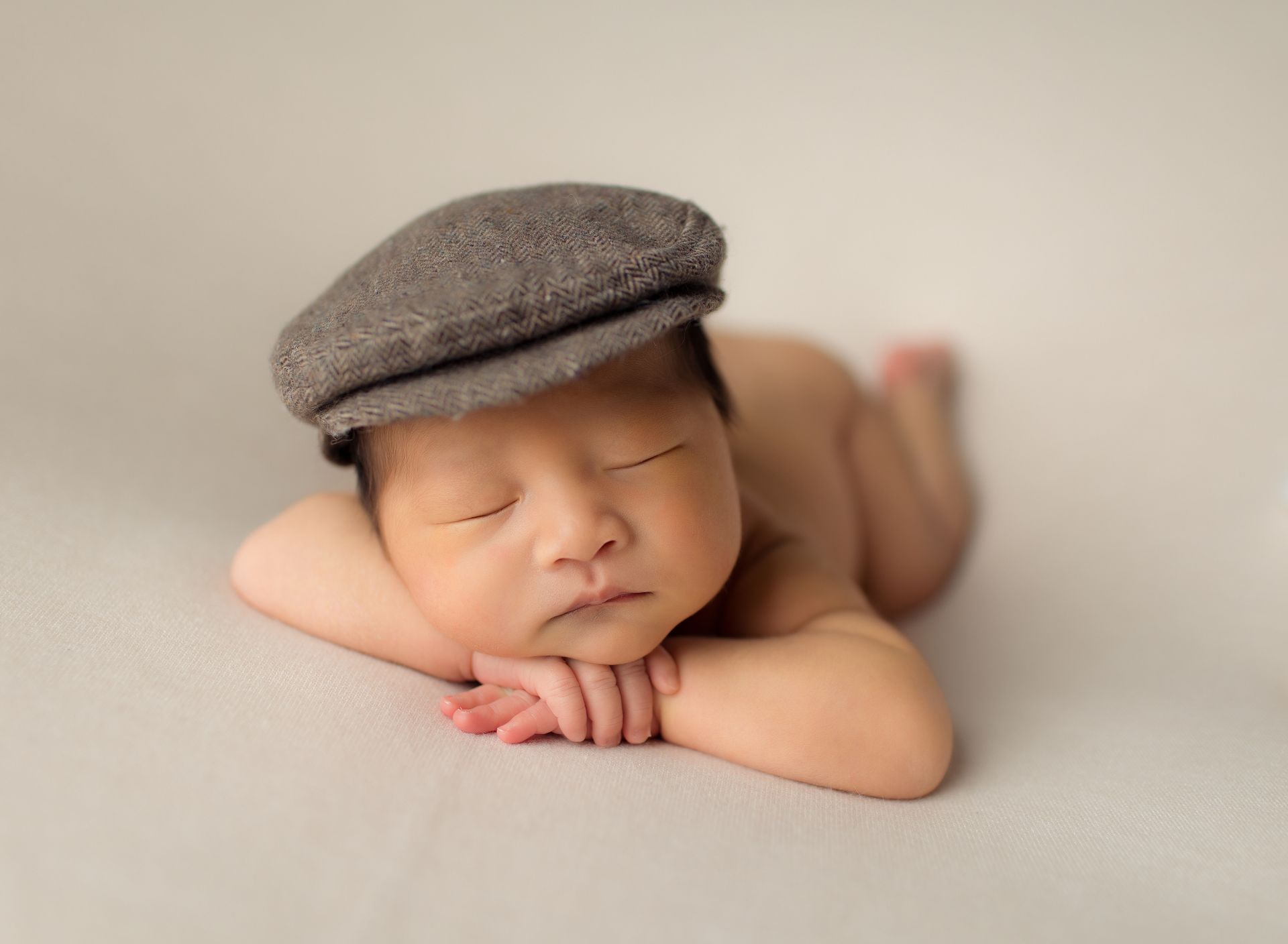 Newborn baby wearing a gray cap, sleeping on its belly with hands supporting its head.