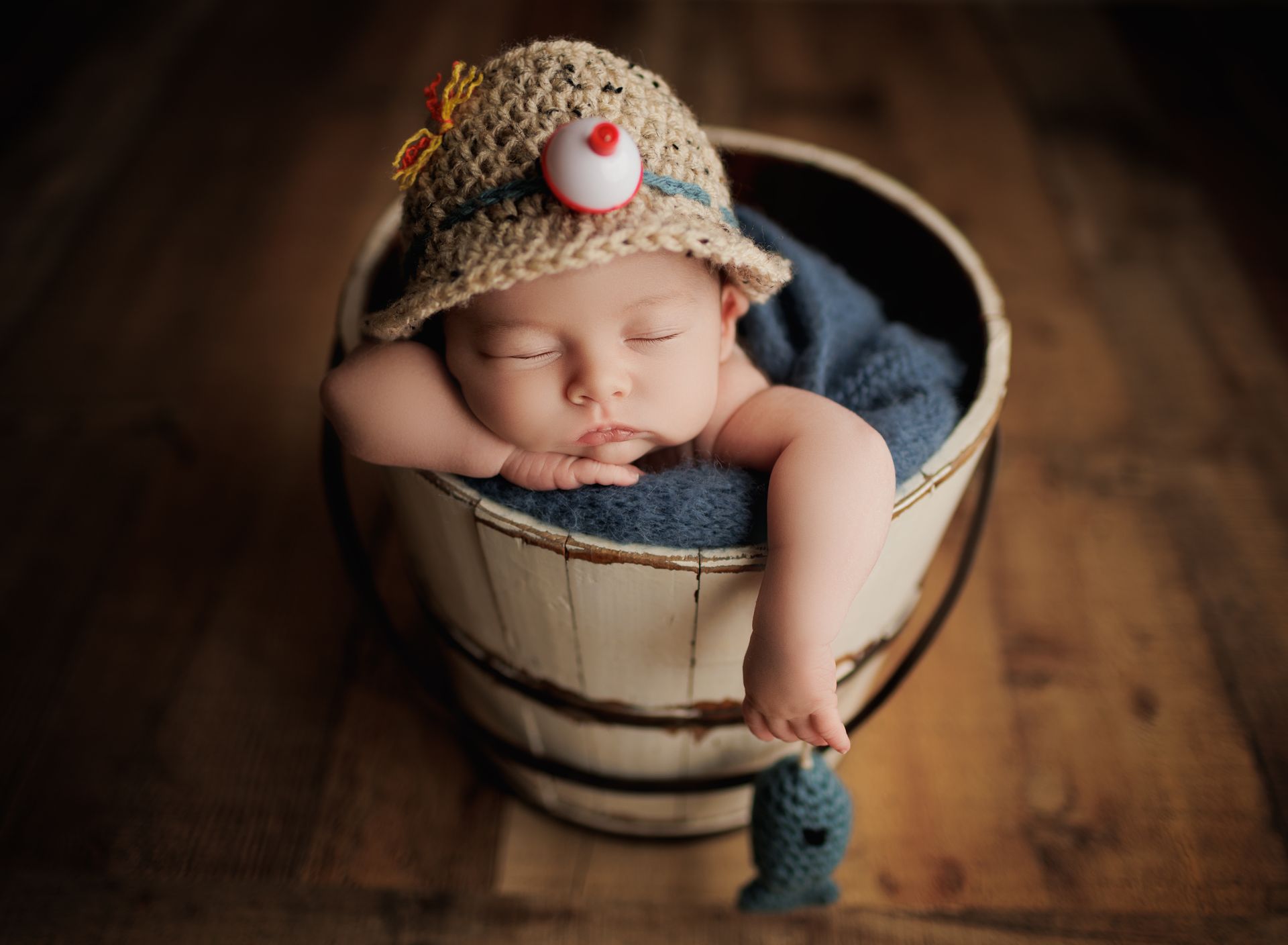 Sleeping baby in a wooden bucket, wearing a fishing hat, nestled on blue fabric.