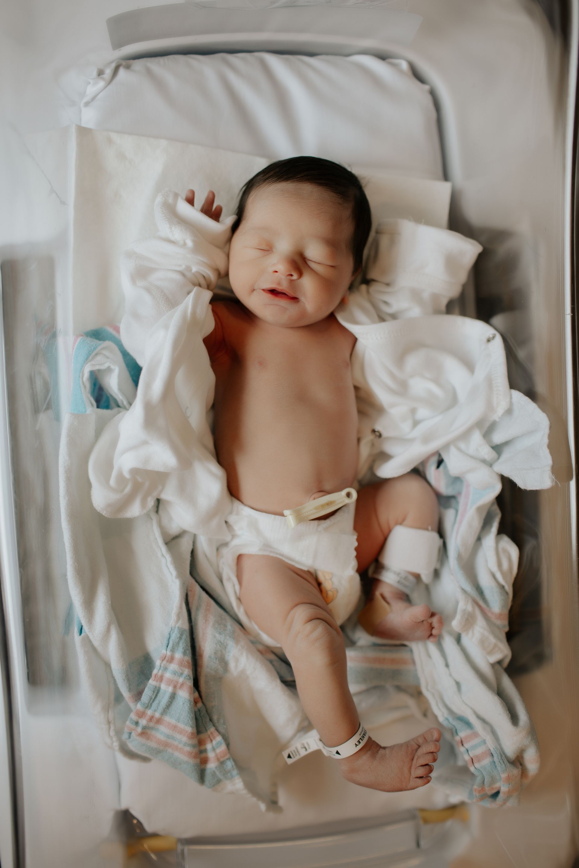 Newborn baby lying in a hospital bassinet, wrapped in a blanket, with wrist and ankle bands.