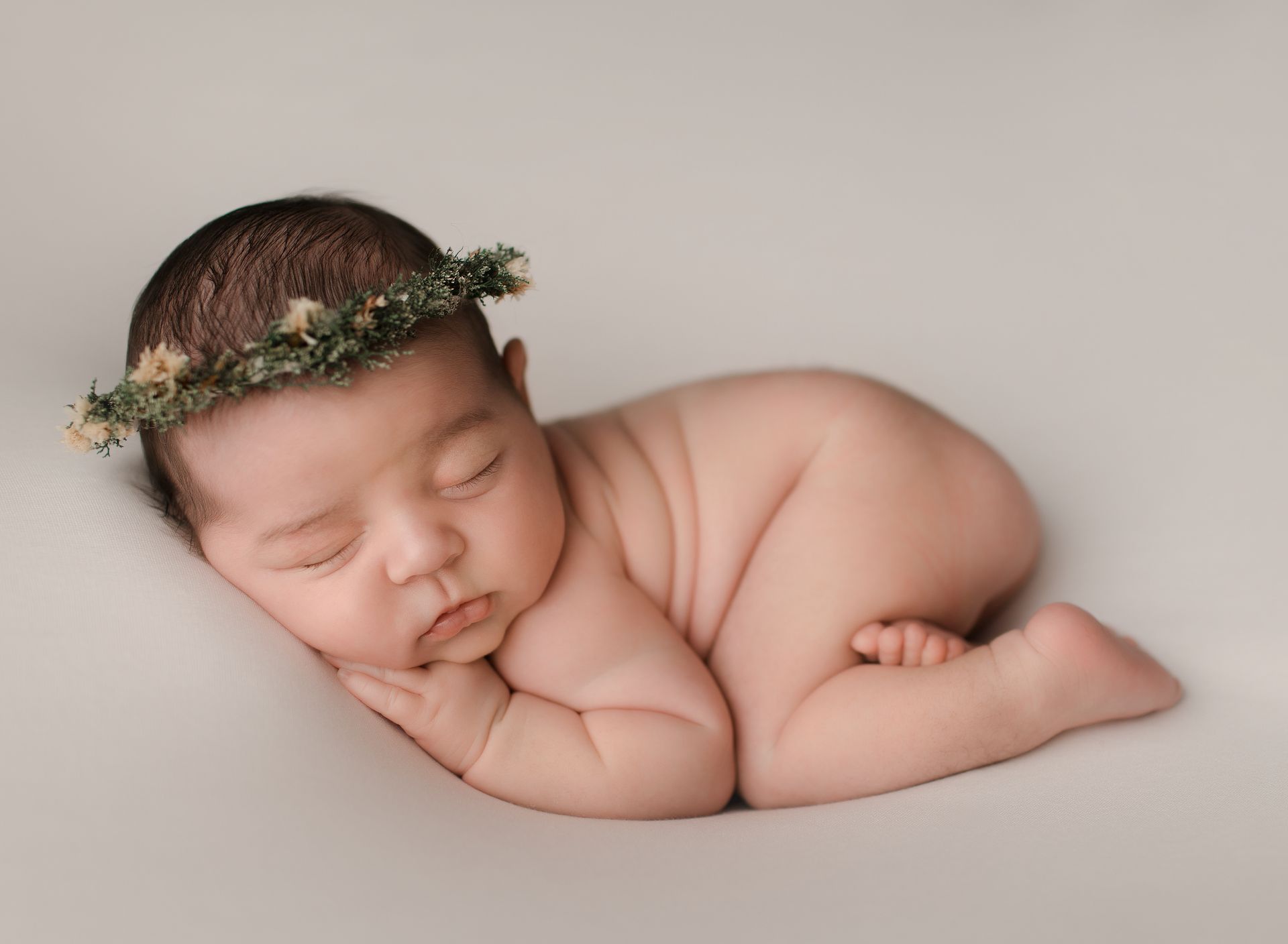 Newborn baby, curled on side, wearing a small floral crown, resting on a neutral-colored surface.