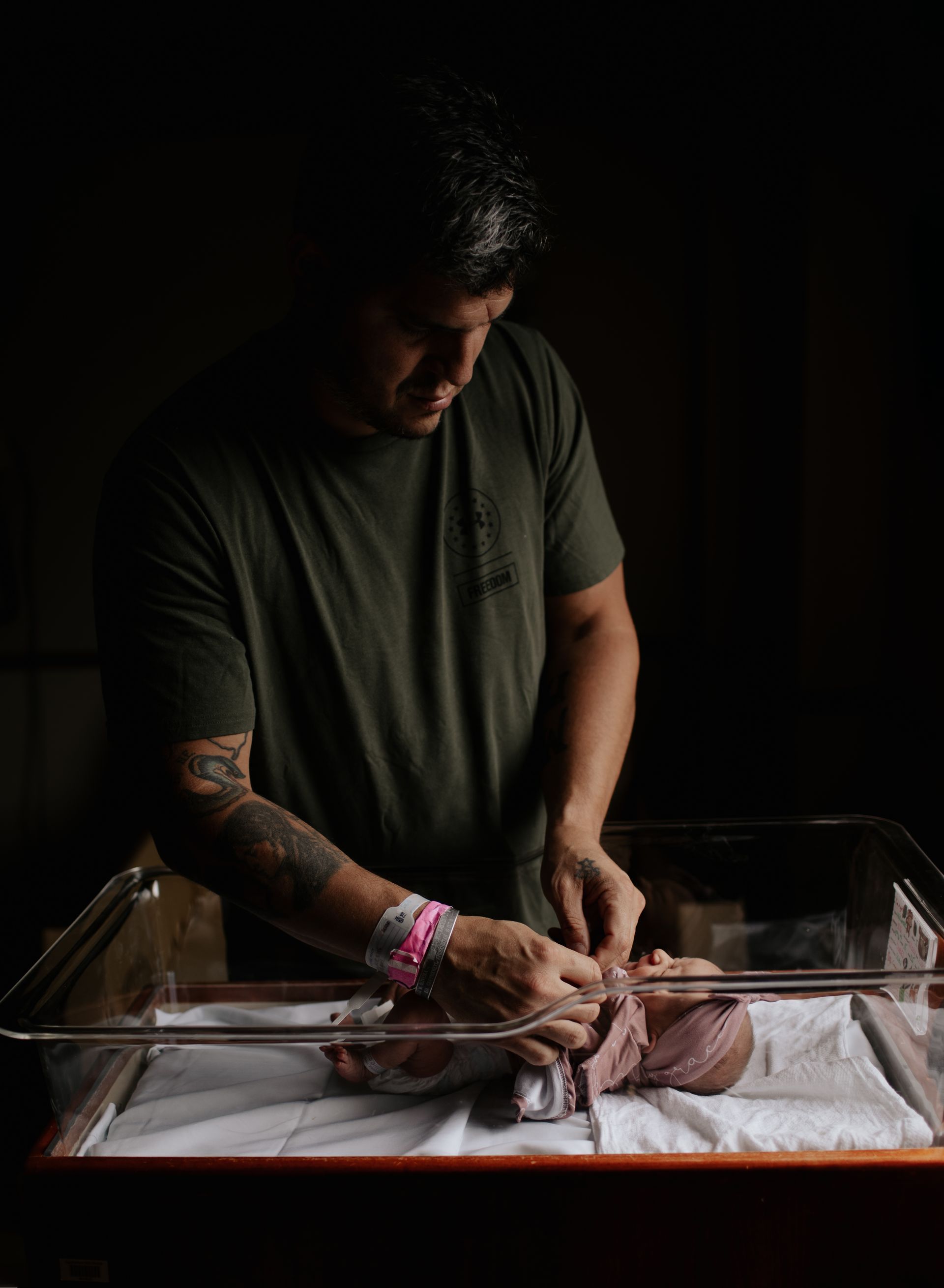 Man in olive shirt, tattoos, cares for newborn in a hospital bassinet. Dark, intimate setting.