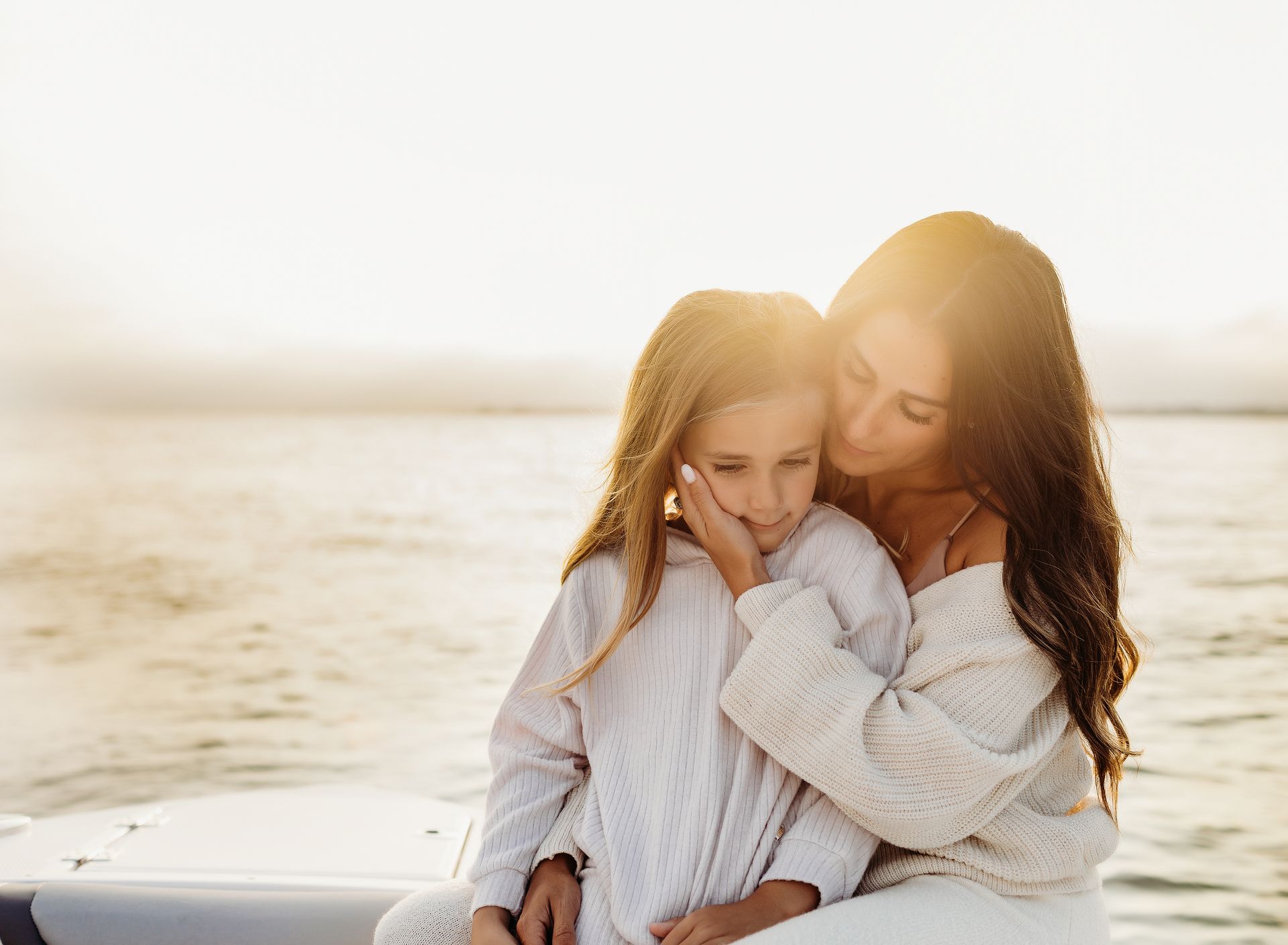 Woman embracing child, both wearing white, by water at sunset.