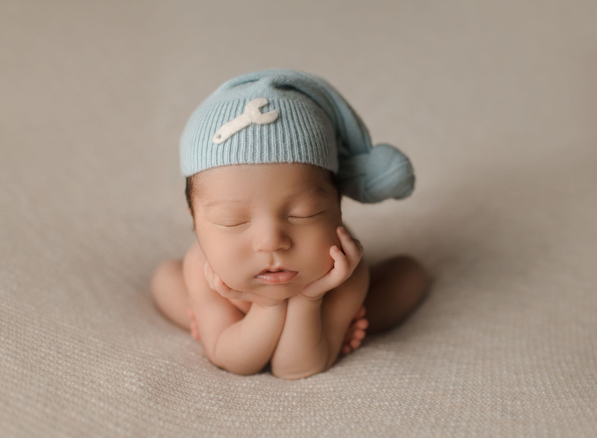 Newborn baby wearing a blue hat with a wrench, sleeping with hands under chin.