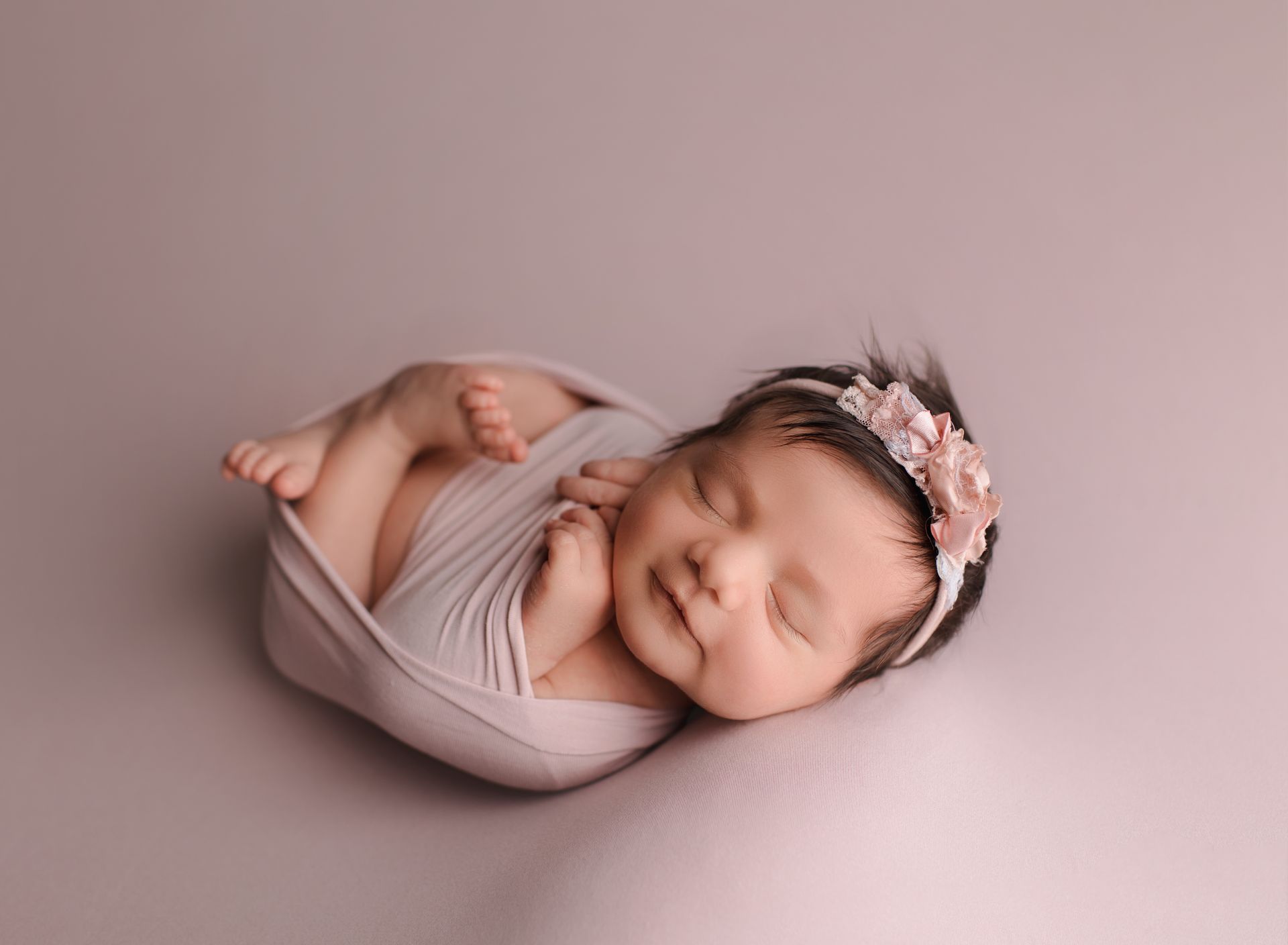 Newborn baby wrapped in pink blanket, smiling with closed eyes, wearing a floral headband.