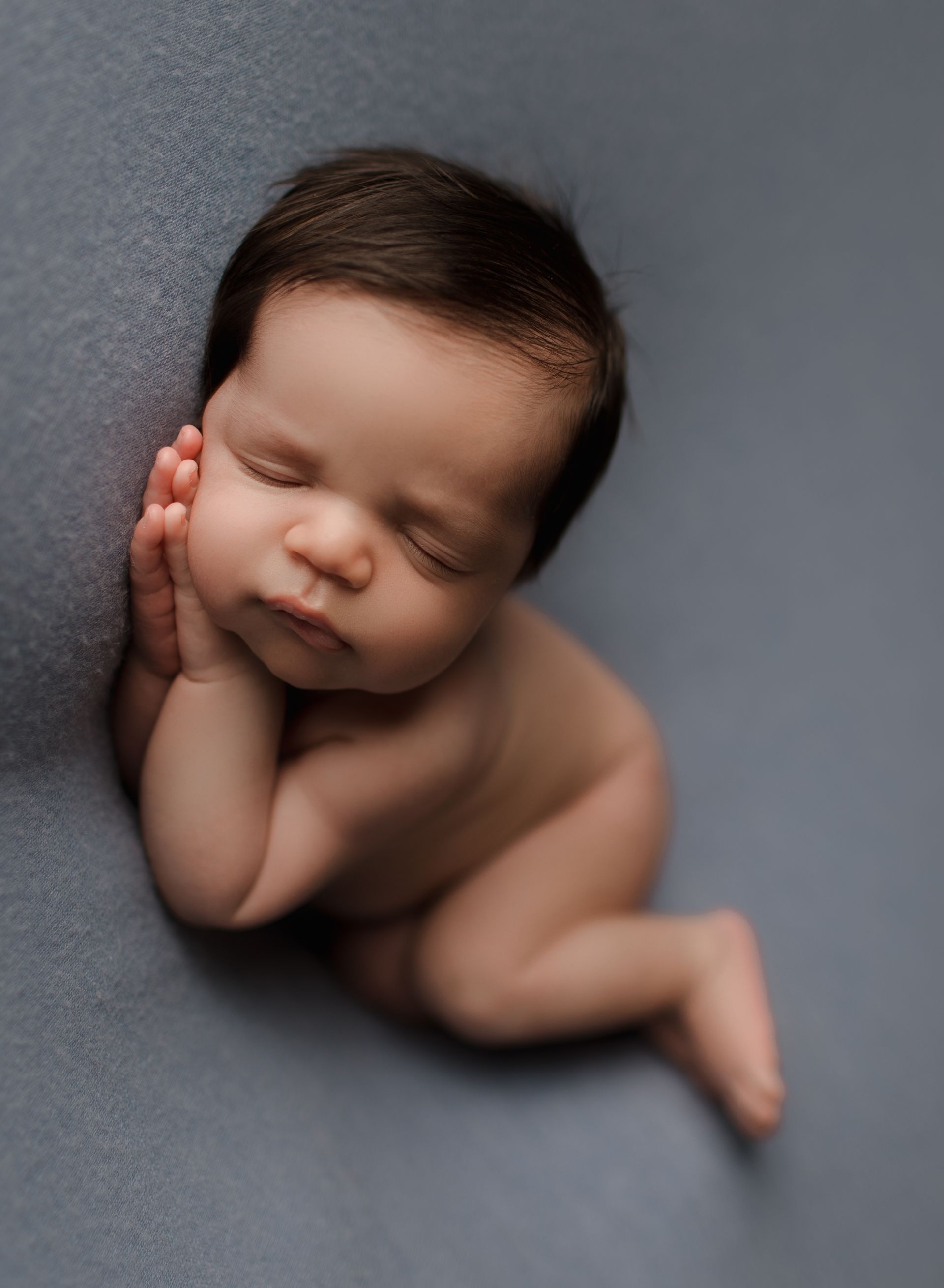Newborn baby, sleeping on a gray surface, hands near face. Brown hair, closed eyes, curled up.