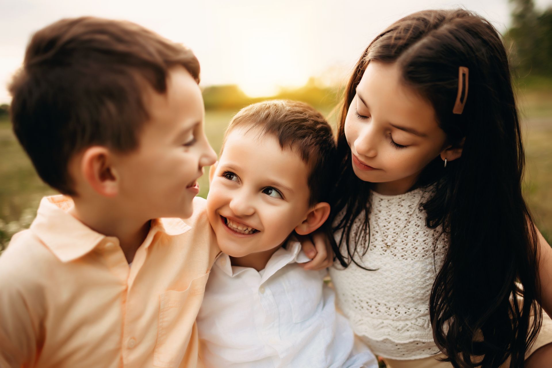 Three children smiling, embracing outdoors. One in the middle smiles widely; others look on with affection.