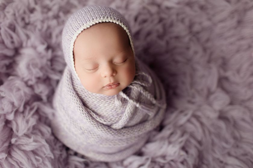 A newborn baby is wrapped in a purple blanket and wearing a hat during a newborn photography session