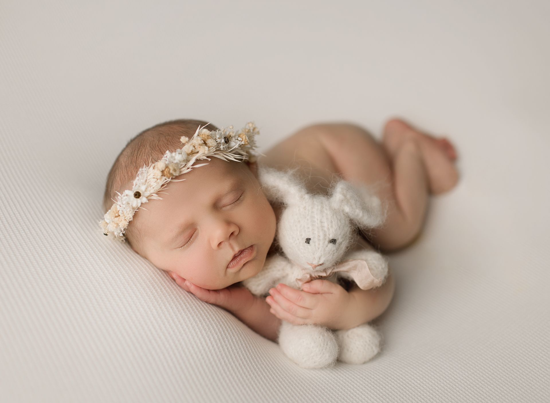 Newborn baby sleeping, wearing floral headband, curled up next to a white stuffed bunny.