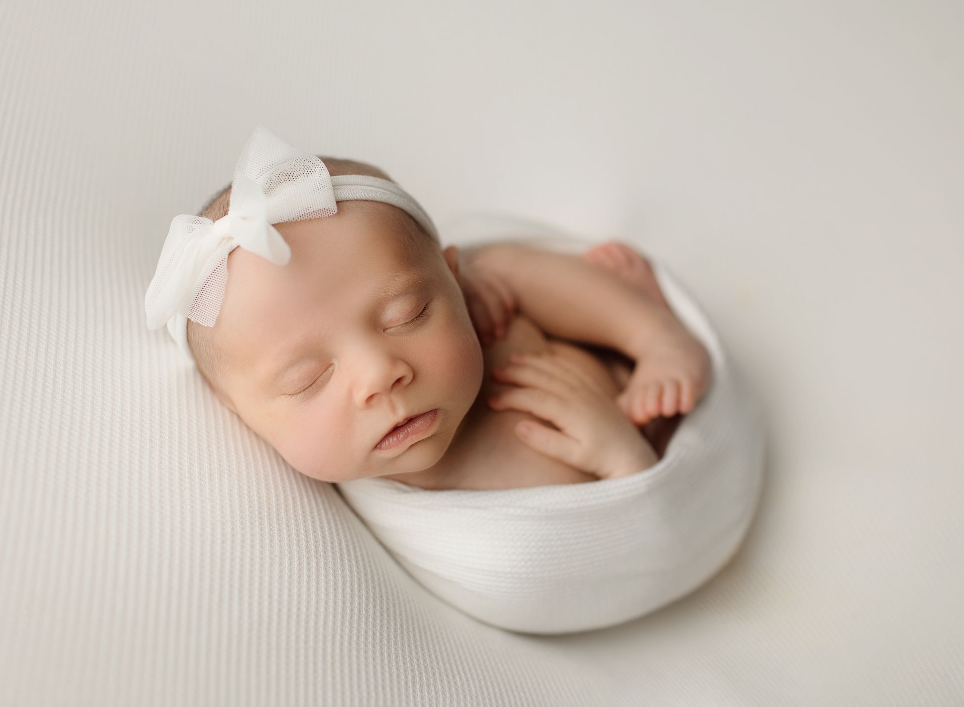 Sleeping newborn baby in white bow headband curled up in a white bowl.