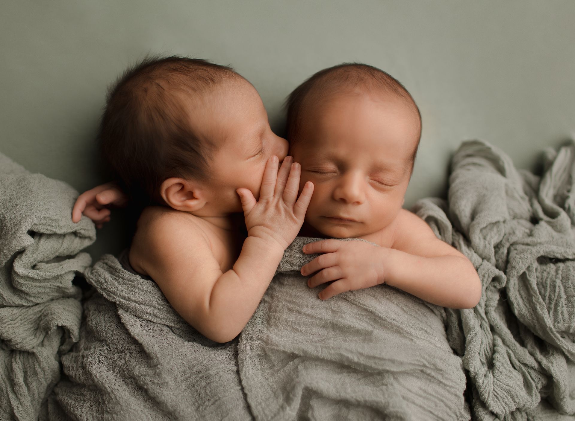 Newborn twins: one whispering in the ear of the other, both wrapped in gray blankets.