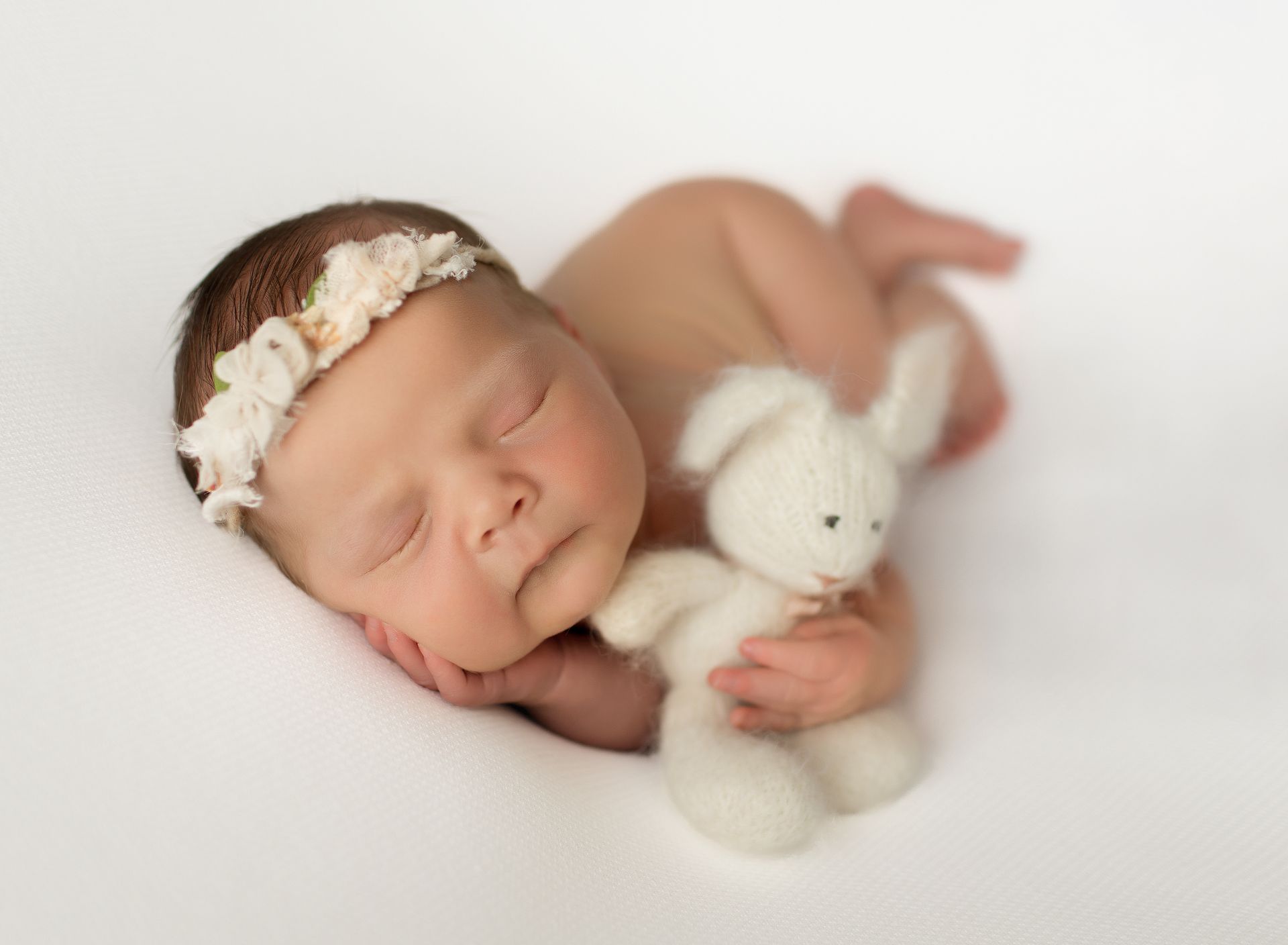 Newborn baby sleeping with a small white stuffed bunny, wearing a floral headband, on a white background.