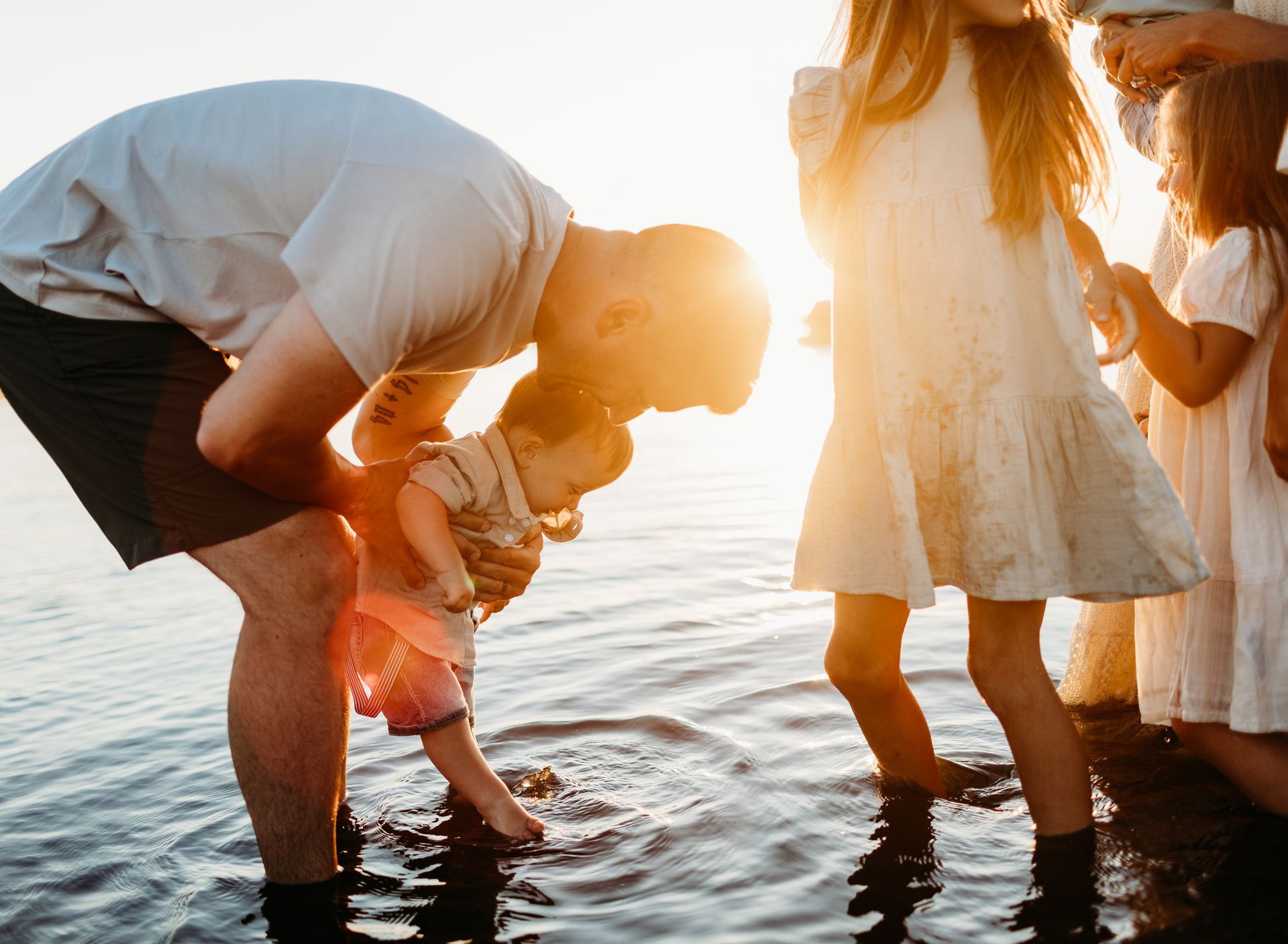 Father playing with toddler in water, sunlit, with two girls nearby.