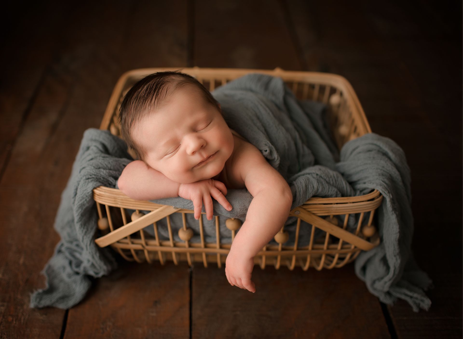 Newborn asleep in a wicker basket, wrapped in blue-gray fabric, on a wooden surface.