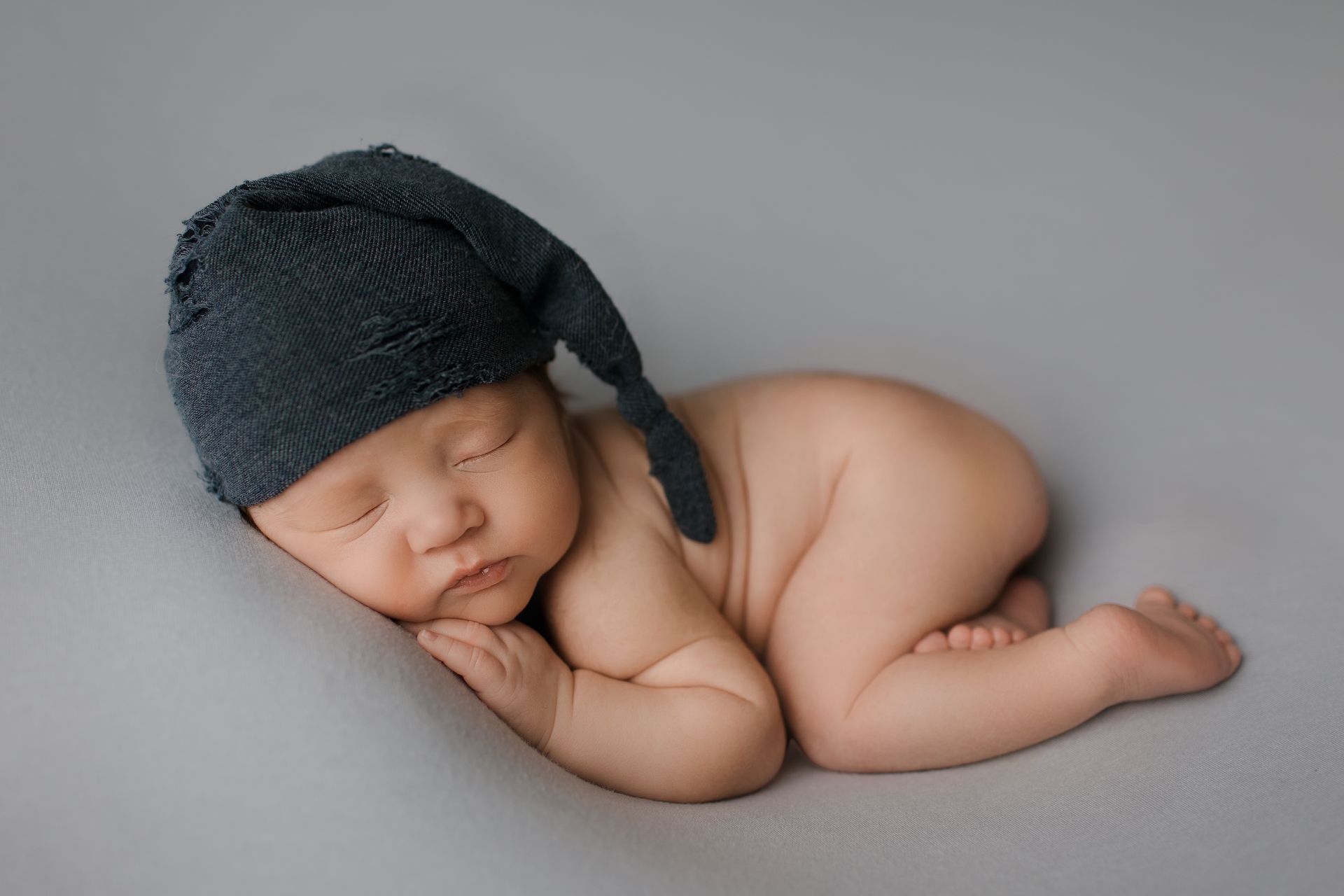 Newborn baby in dark blue hat, curled up sleeping on grey backdrop.