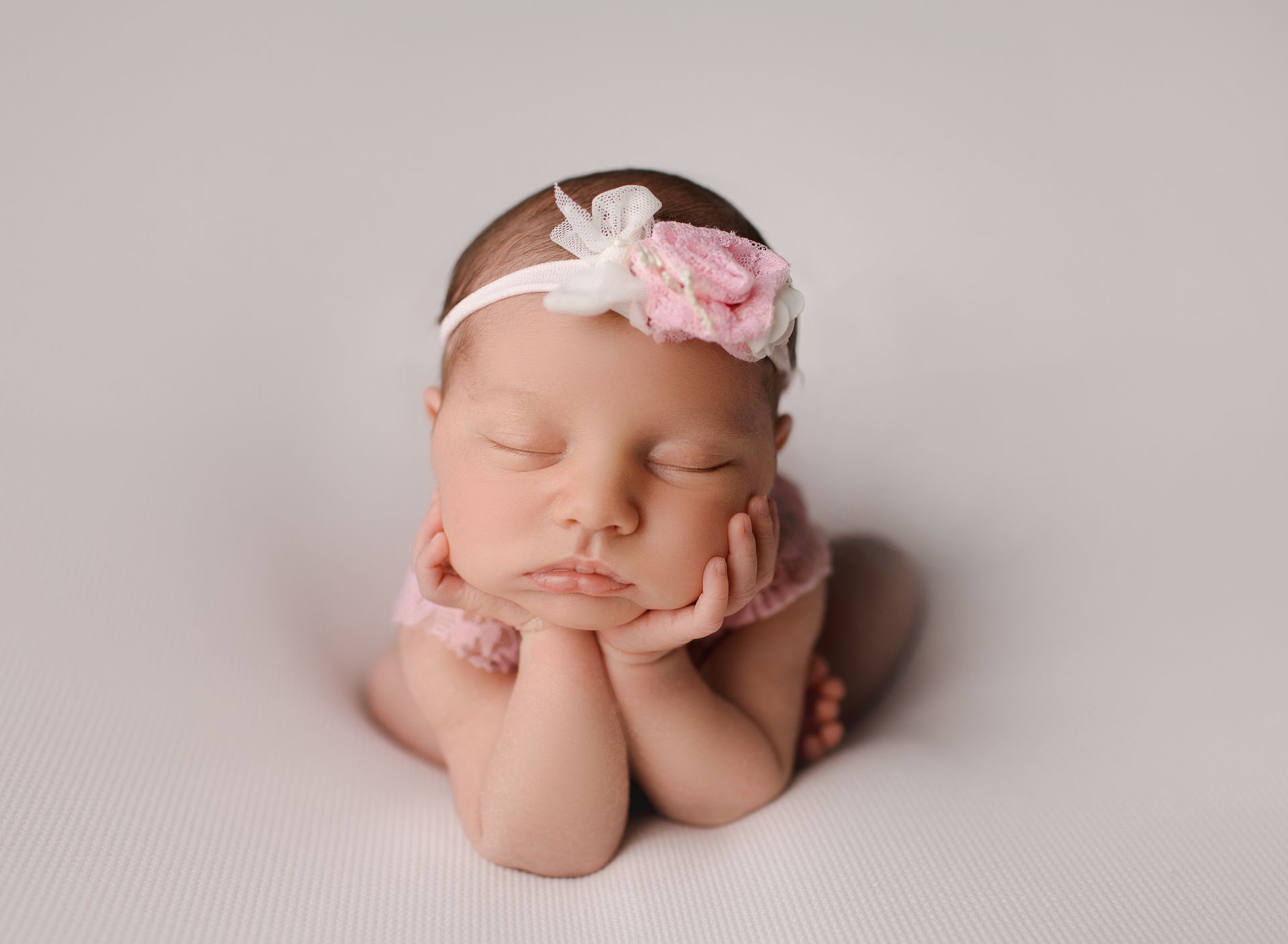 Newborn baby with eyes closed, wearing pink flower headband, rests chin on hands, on white surface.