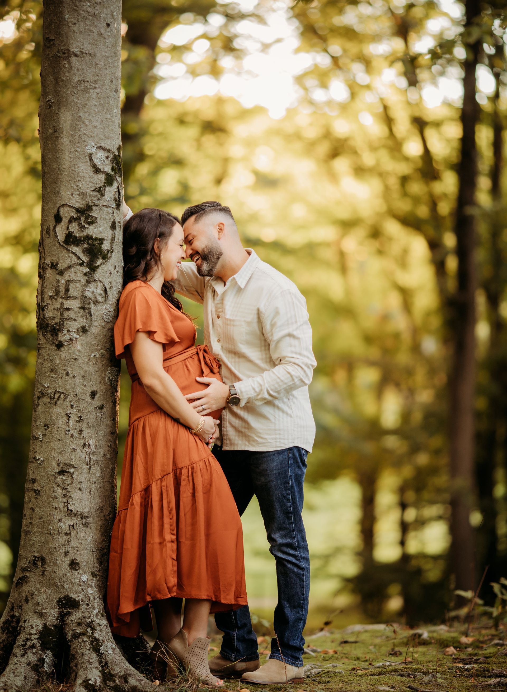 Pregnant couple embraces beside a tree in a sunlit forest. Woman in orange dress, man in a button-down shirt.