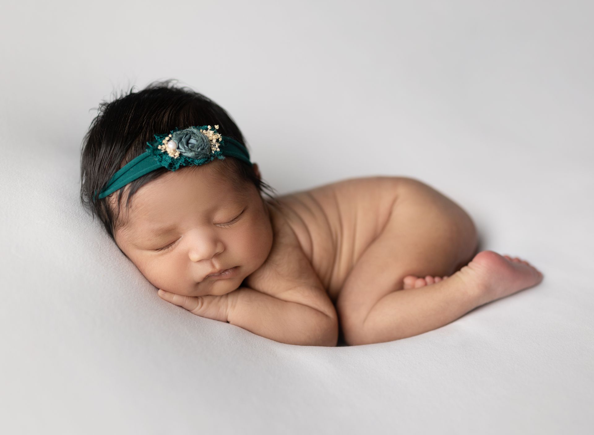 Newborn baby asleep on a white surface, wearing a teal headband with floral detail.