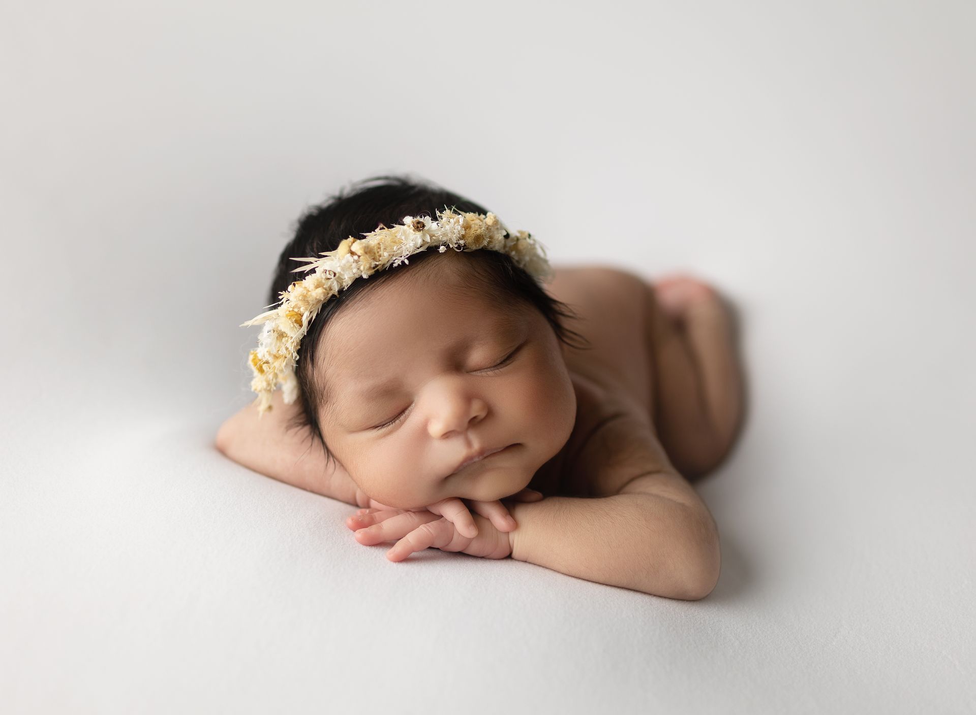 Newborn baby, dark hair, wearing a floral headband, resting on arms, on white backdrop.