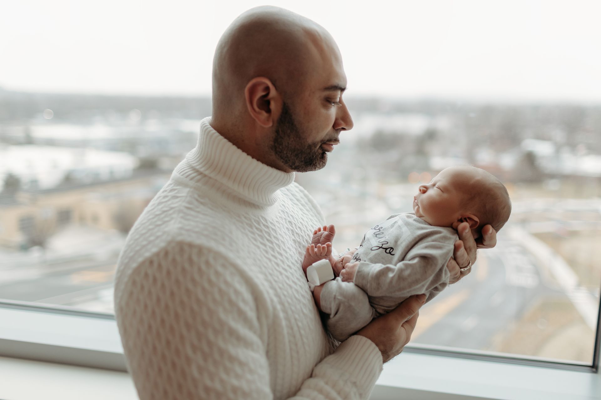 Man holding a newborn baby by a window; both are dressed in white.
