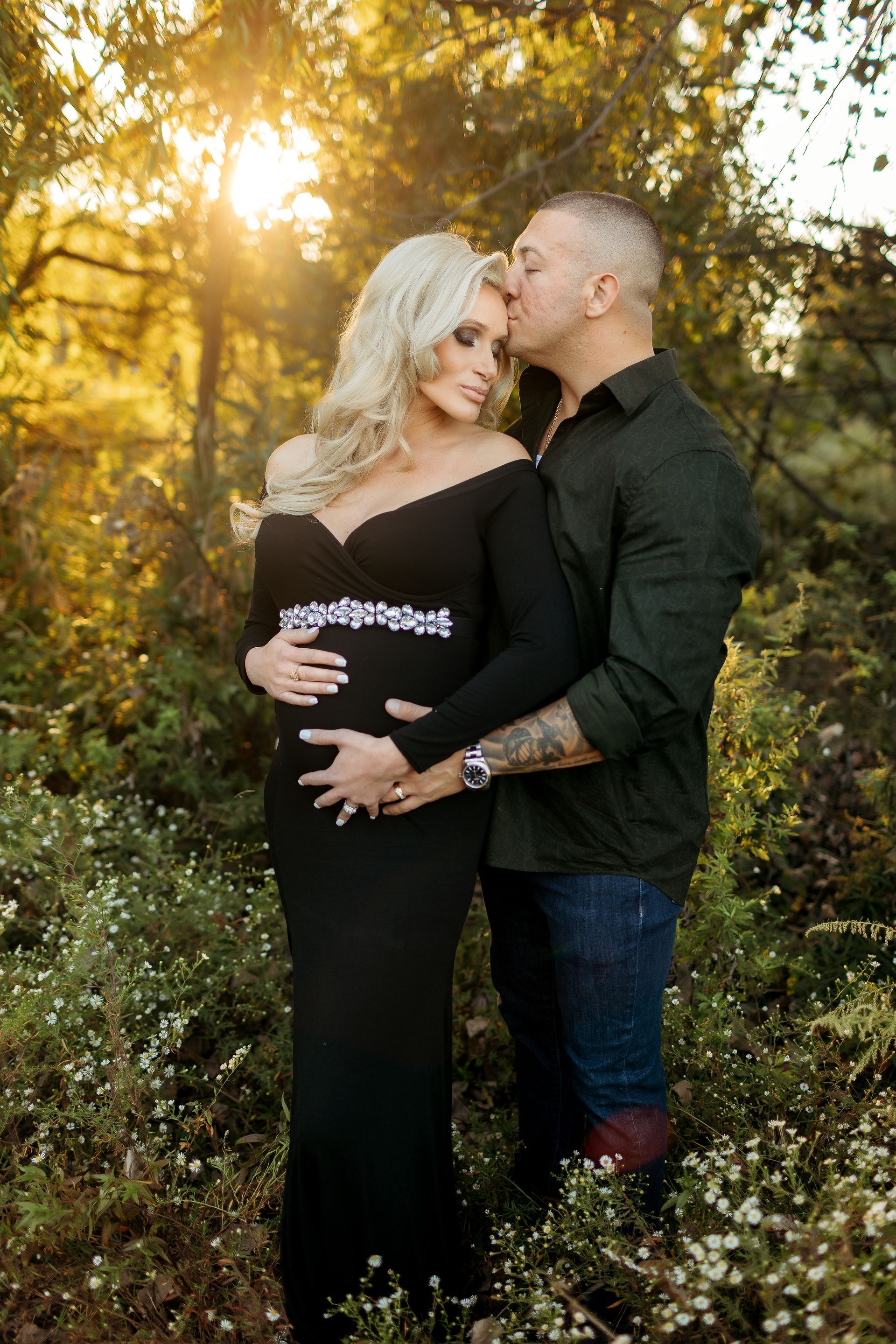 A man is kissing a pregnant woman on the cheek in the woods.