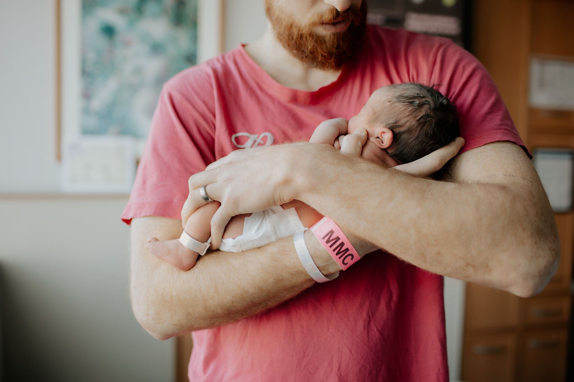 Man in pink shirt holding newborn, both wearing hospital bands.