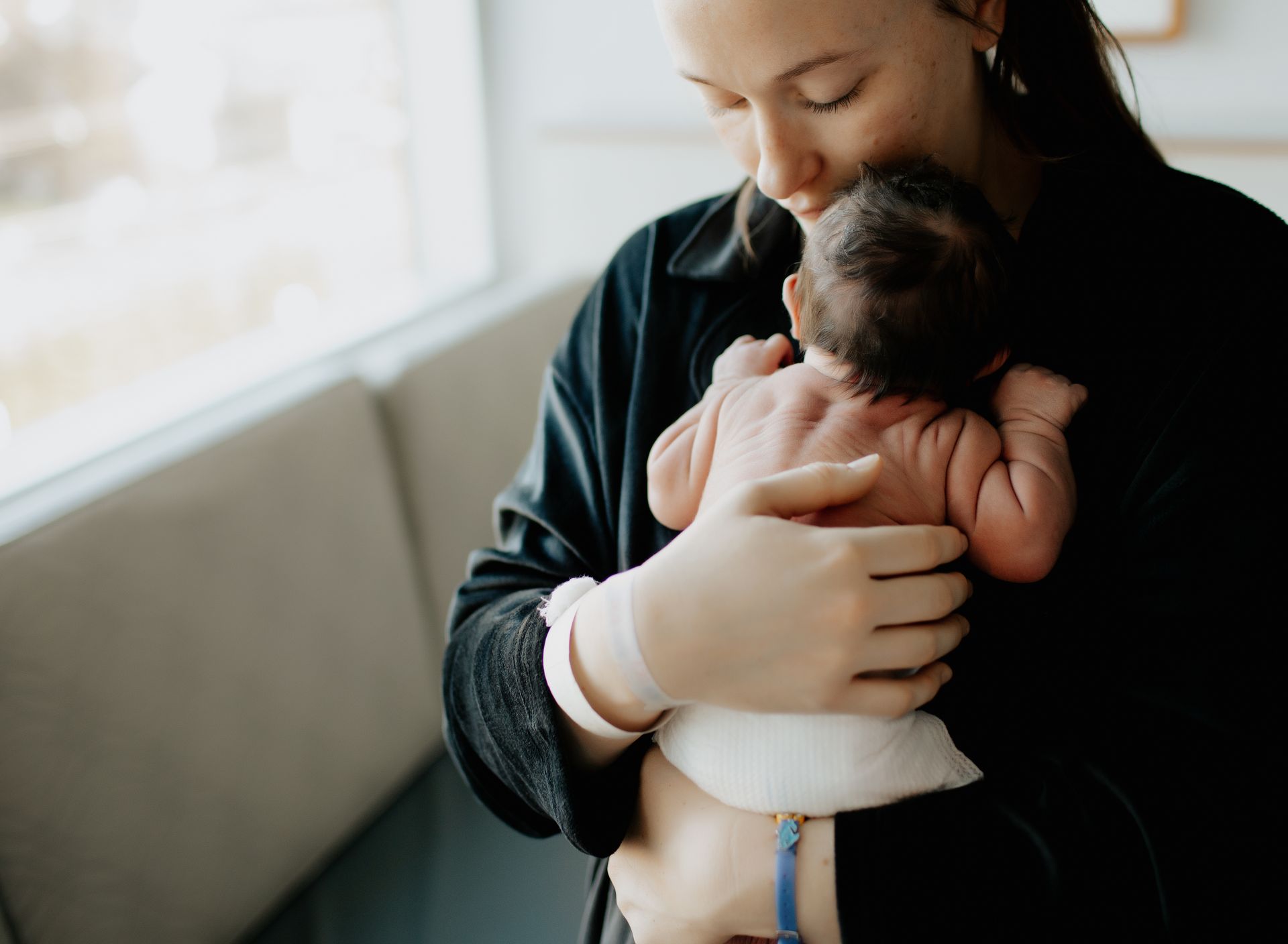Woman holding newborn baby, head touching the baby's head. Baby is wearing a diaper, indoors near a window.