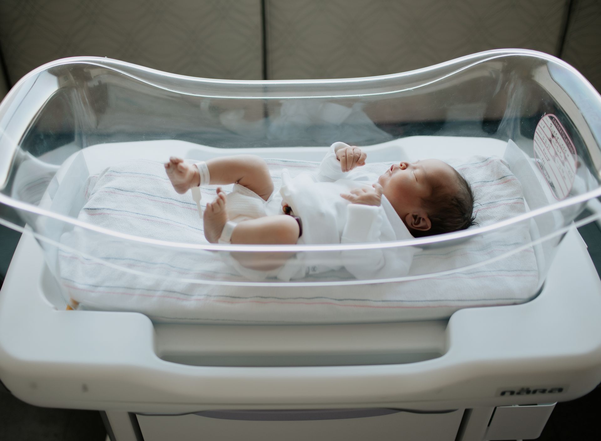 Newborn baby in clear bassinet on a white hospital stand.