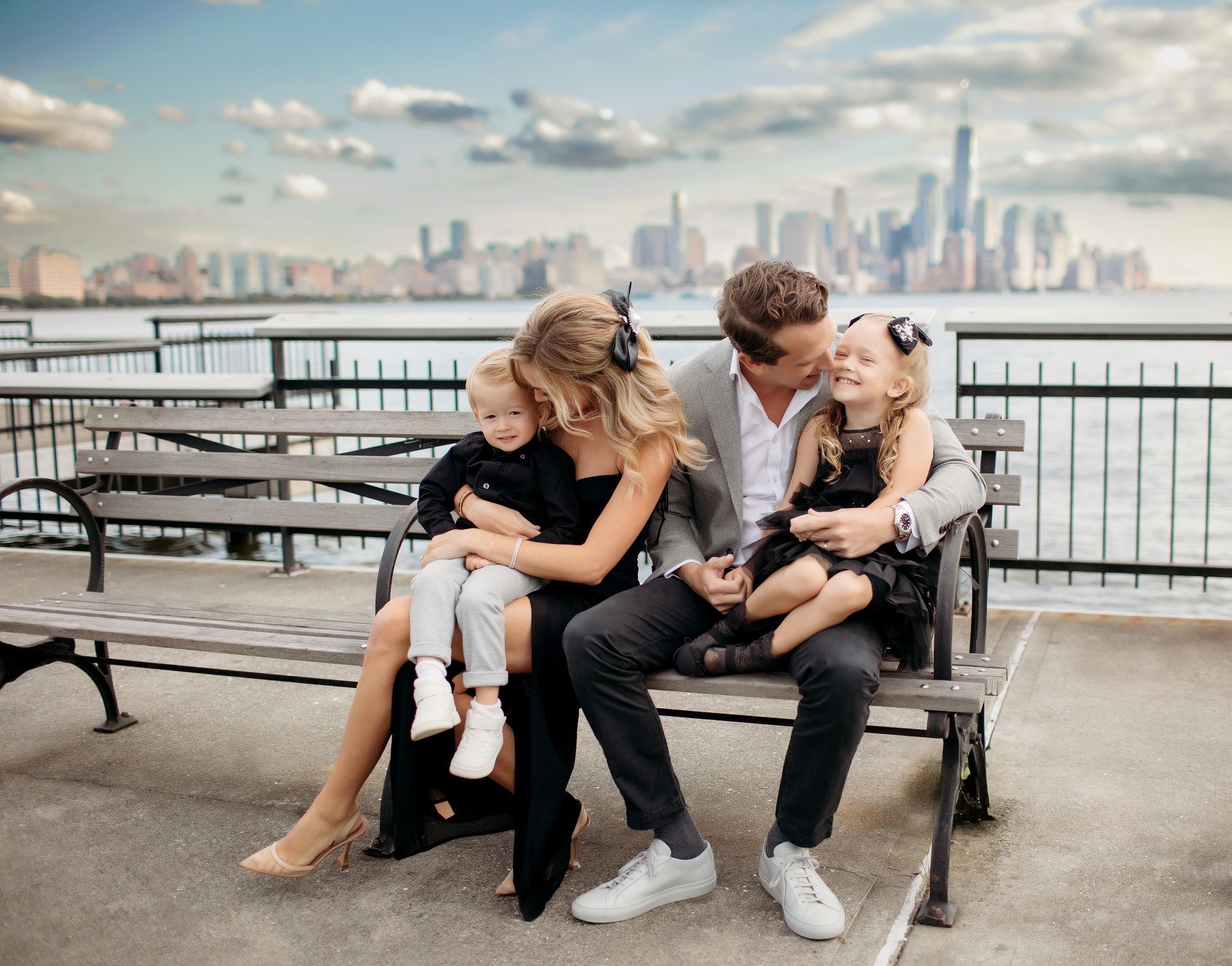 Family of four sitting on a bench; NYC skyline in background. Parents embrace children; sunny day.
