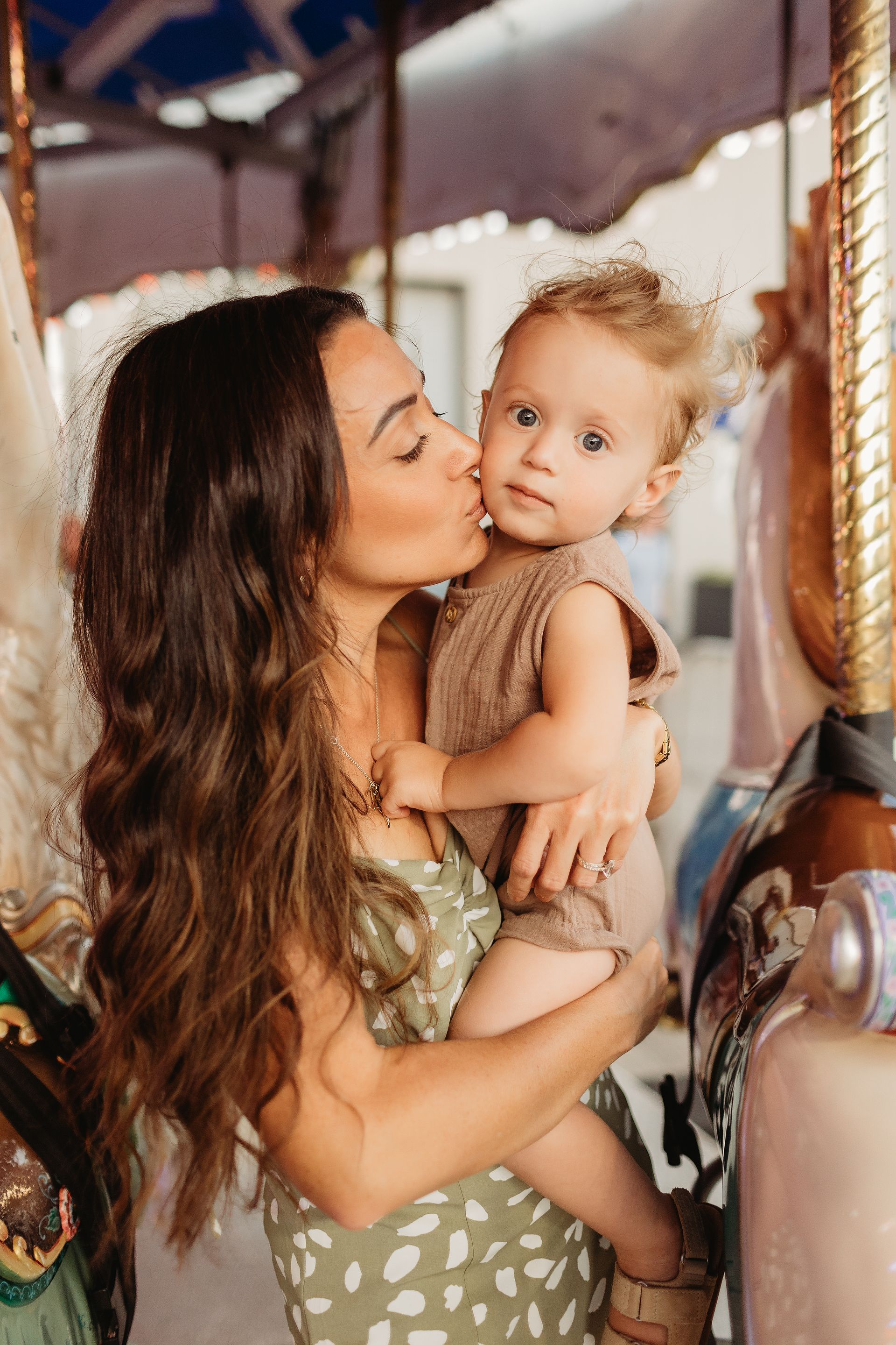 Woman kissing a child on the cheek near a carousel. They are both wearing light-colored clothes.