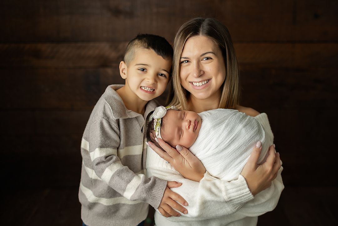 Woman holding newborn with older child, all smiling. Brown wood background.